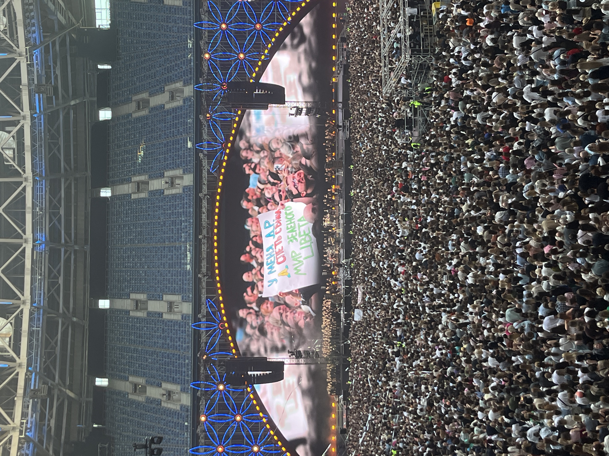 The image captures a large outdoor concert event at the Gazprom Arena in Saint Petersburg, Russia. A massive crowd of spectators fills the stadium, extending from the stage area far into the stands. The main stage features a large screen displaying a close-up of audience members holding up a white sign with text written in Cyrillic. Decorative blue floral light installations with yellow accents are prominent along the top of the stage. The stadium seating, primarily blue, is visible behind the screen and above the crowd. The lighting suggests it is evening or nighttime.