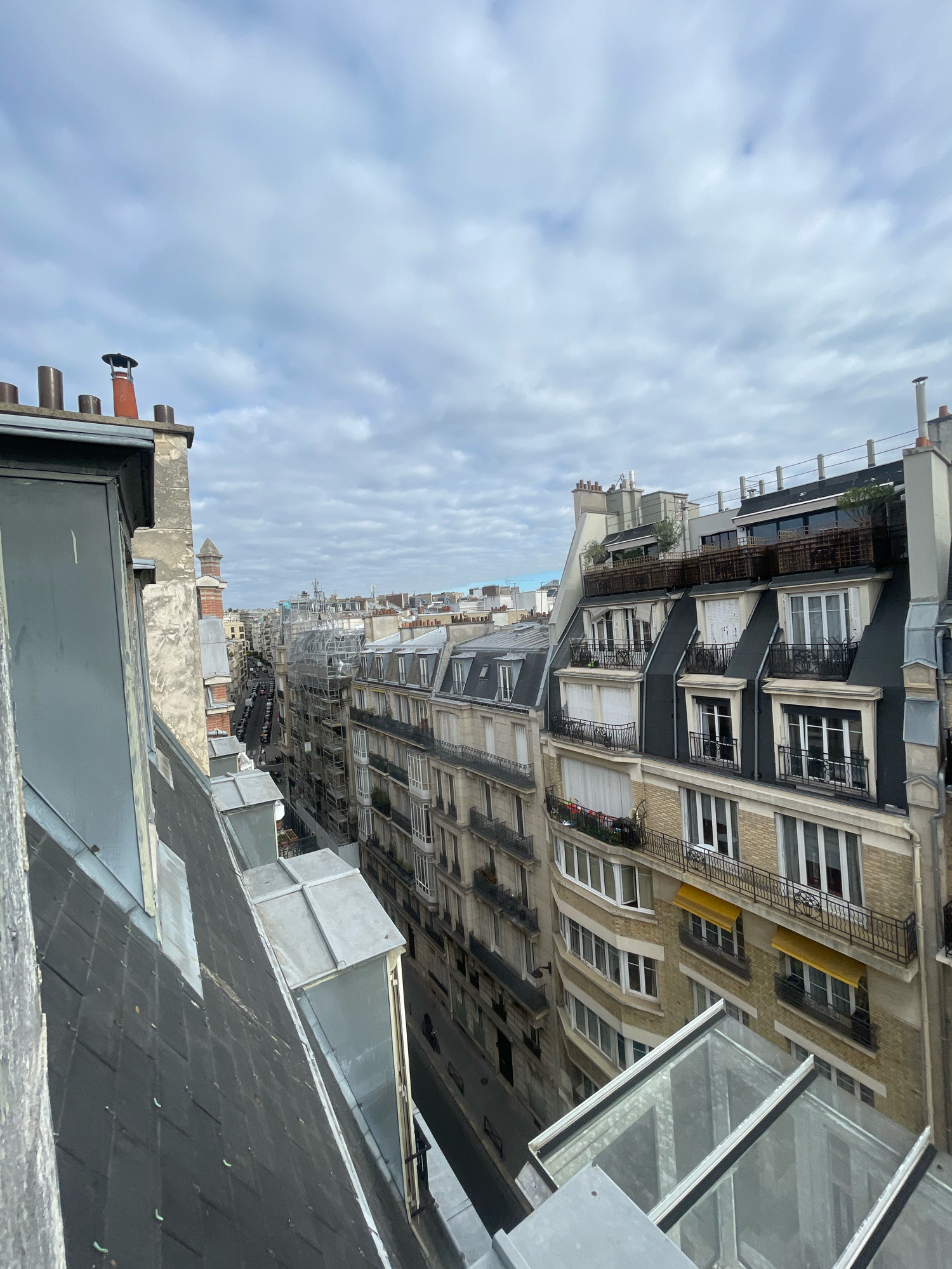 This is a high-angle, eye-level, medium shot of a street in Paris, France, on an overcast day. The scene is dominated by the architecture of Parisian buildings, with their distinctive mansard roofs and narrow facades. In the foreground, the slanted, dark gray roof of a building is visible, with several metal skylights and a gray painted wooden railing. Further back, a row of buildings stretches down a street lined with parked cars. The buildings have multiple stories, with numerous windows, many featuring black wrought-iron balconies. Some buildings are undergoing renovation, with scaffolding visible. Yellow awnings are visible on some of the lower windows. Chimneys protrude from various rooftops. The sky is filled with gray clouds. No people are clearly visible in the scene.