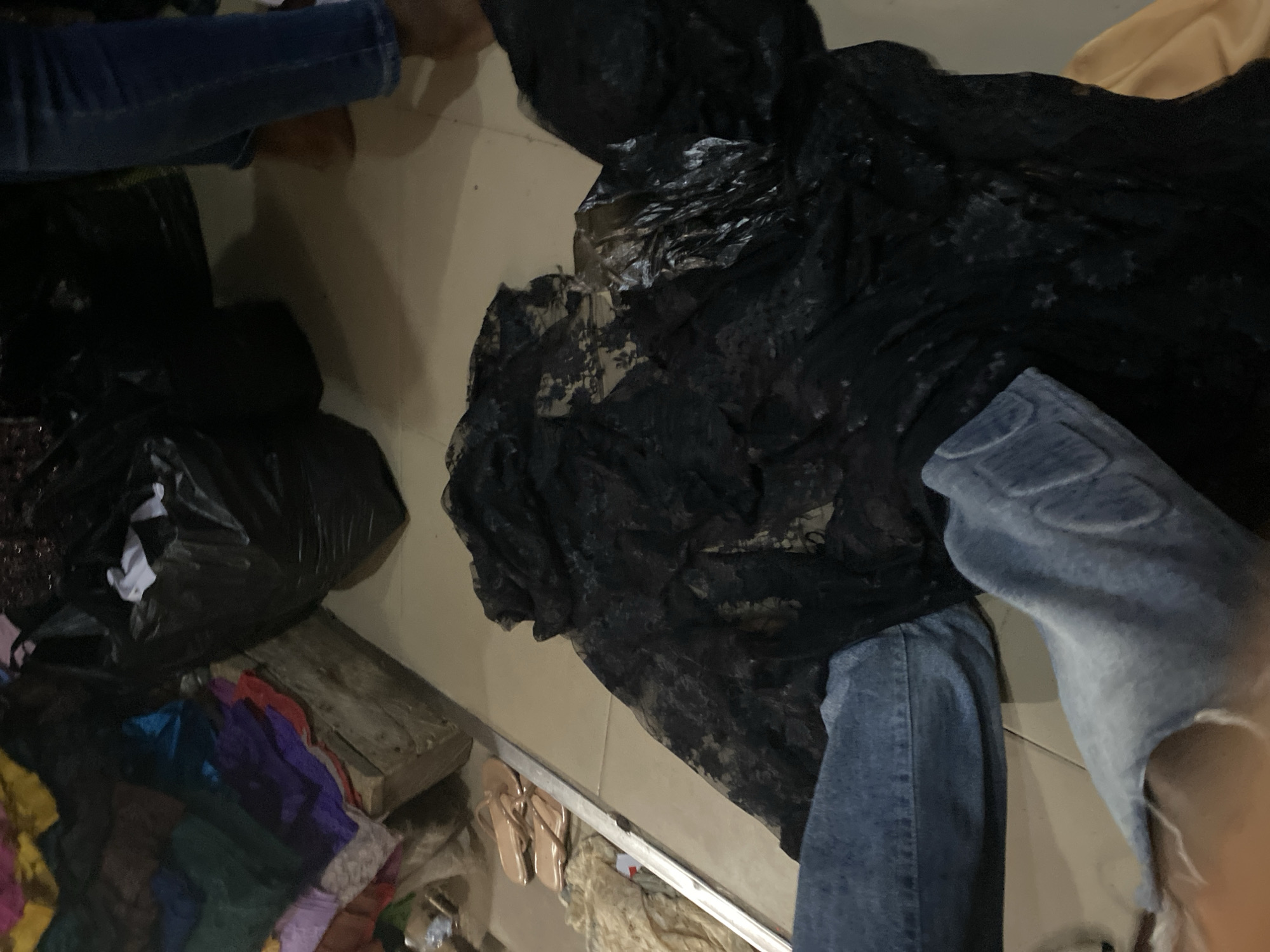 A person's legs, clad in distressed blue denim jeans, are visible in the foreground of the image, with a torn section revealing skin. A large piece of black, sheer fabric, possibly lace or net, is draped over the floor in front of them and extends out of frame to the right. To the left, a metallic railing runs along a surface holding a pair of light brown flip-flops and some beige fabric. In the background, to the left, is a stack of colorful fabrics, including green, purple, and red. Behind the fabrics, a large black trash bag is visible, and a portion of another black trash bag is seen to the right, partially obscuring a person's legs in blue jeans and brown footwear. The floor appears to be tiled in a light beige color. The scene suggests a marketplace or stall in Jalingo, Nigeria, likely focused on textiles or clothing.