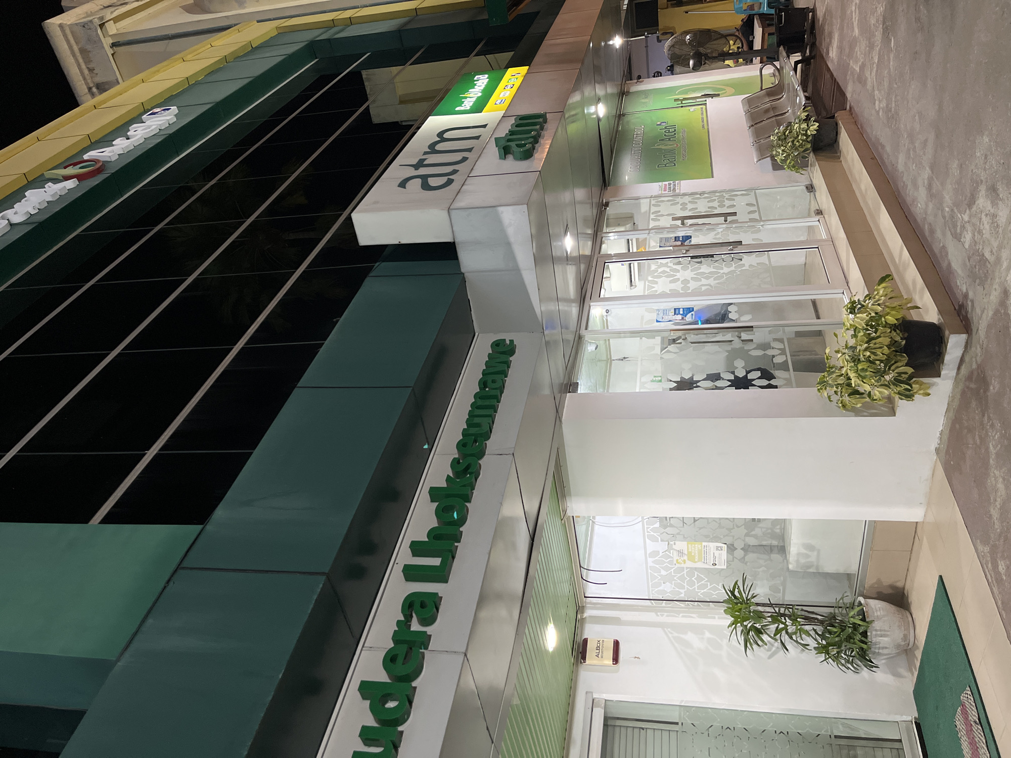 The image depicts the exterior of a Bank Aceh branch, identified by prominent signage reading "Bank Aceh" and "atm." The building features a green and dark glass facade, with some yellow accents. The entrance area is partially visible, with glass doors and a small set of stairs leading up to them. Inside, an ATM machine is visible. Several potted plants are placed outside the entrance. A sign with "SECURITY CONTROL" is also visible. The scene appears to be in a city, and the banking institution suggests it is likely in a commercial or urban area of Lhokseumawe, Indonesia. The lighting indicates it is nighttime or dusk. There are no people clearly visible in the scene.