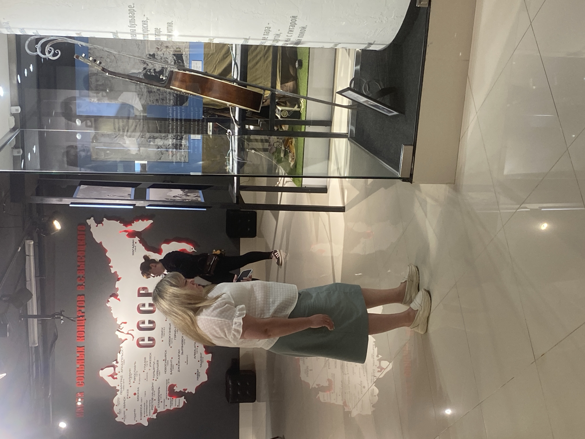 A woman with blonde hair is looking at a display case that contains a guitar. She is wearing a white blouse and a green skirt. In the background, there is a map of the USSR. Another woman in a black jacket is standing next to her, looking in the same direction. The scene is indoors, likely a museum. The text on the wall above the map of the USSR appears to be in Russian. The display case also has text on it. The floor is tiled and reflective.