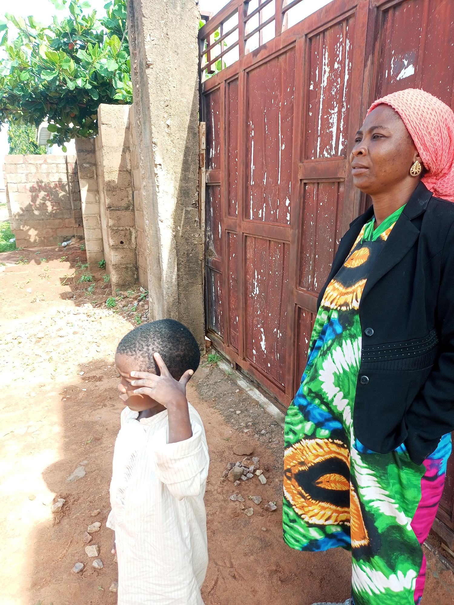 In the city of Tempure, Nigeria, a woman wearing a pink headscarf and a black blazer stands by a rustic, weathered red metal gate. She is looking off to the right, her expression thoughtful. Beneath her blazer, she wears a vibrantly patterned green and orange garment. To her left, a young boy in a light-colored striped shirt stands with his hand raised to his forehead, shielding his eyes from the sun. The ground is dry and dusty, with scattered stones. Behind them, a rough concrete wall runs alongside a section of a brick wall, partially obscured by lush green foliage from a tree overhead.