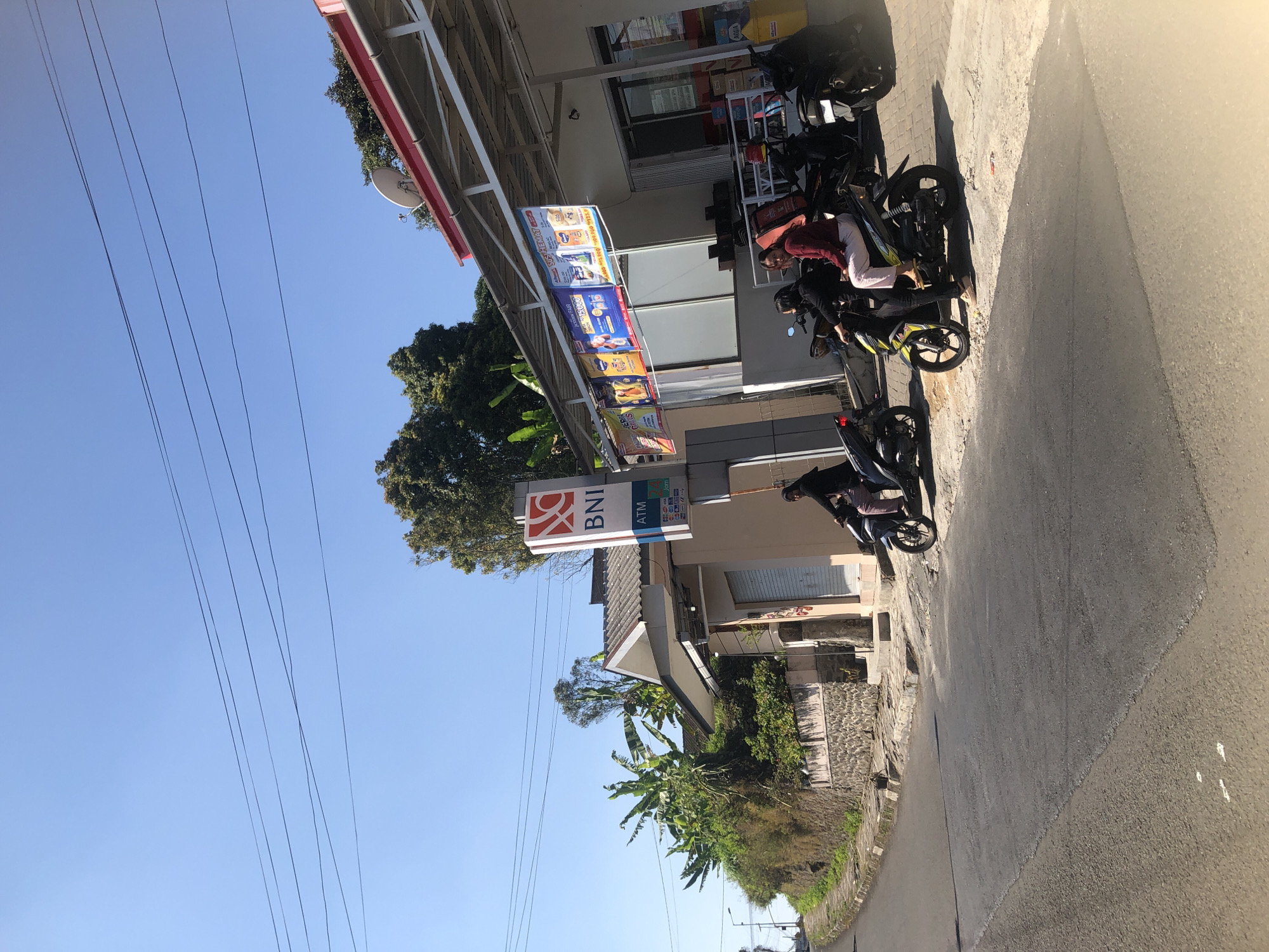 This is a sunny daytime, eye-level, medium shot of a street scene in Trawas, Indonesia. The image captures a section of a street with a slight incline, flanked by a building on the right and a stone wall with vegetation on the left. The building appears to be a commercial establishment, possibly a convenience store, with a red-trimmed canopy extending over the sidewalk. Banners advertising various products are hung along the canopy. A BNI ATM sign is prominently displayed on the side of the building.
Several motorcycles are parked and in use on the sidewalk in front of the building. Two women are seated on a motorcycle in the foreground, with one appearing to operate the vehicle. Another motorcycle is partially visible to the left, and two more are further to the right. The road surface is asphalt, with some visible markings and a curb along the edge. Lush green vegetation, including banana trees, grows on the hillside to the left. Power lines are strung across the clear blue sky. The overall impression is a typical street view in a tropical urban or semi-urban environment.