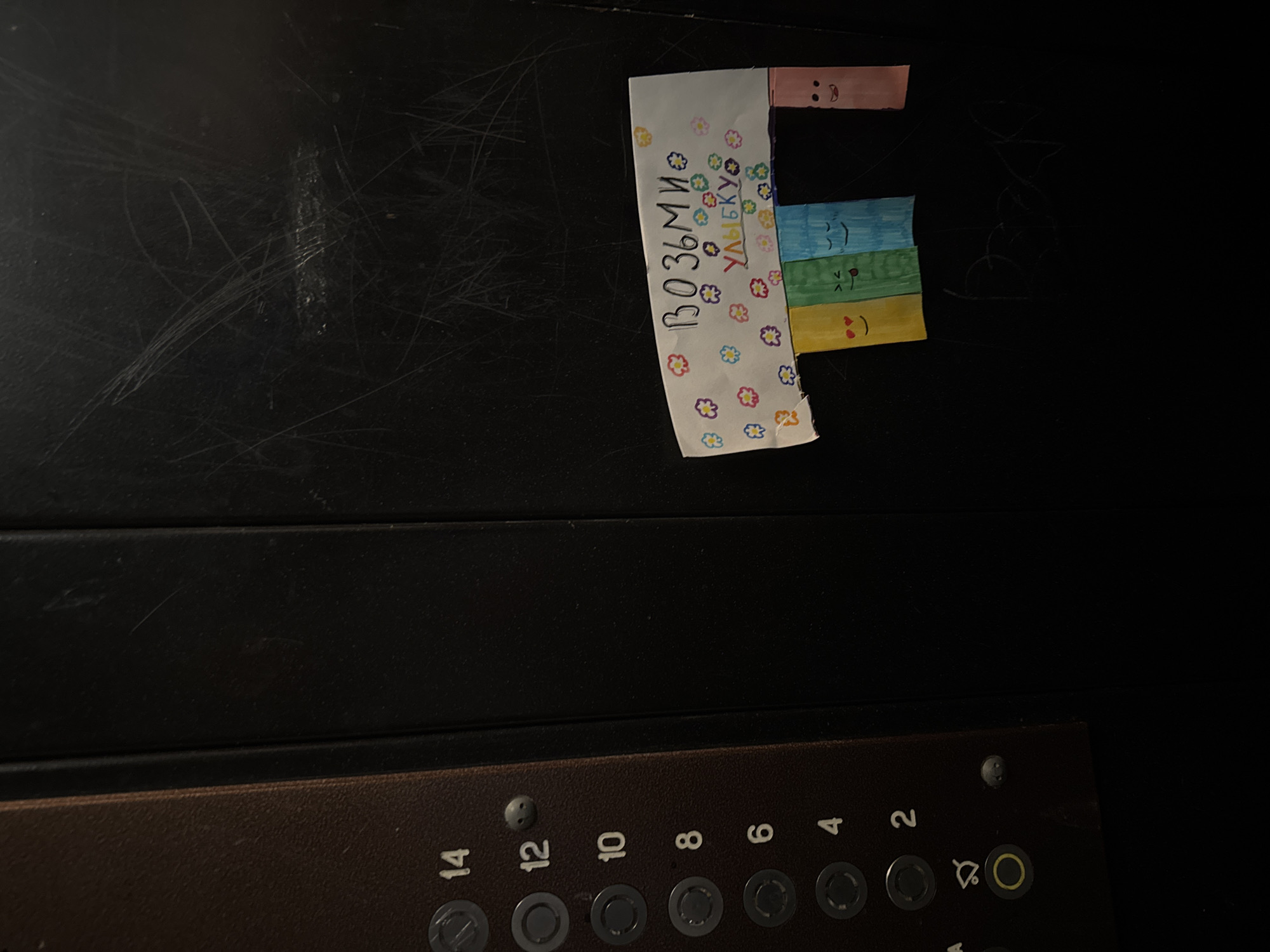 A close-up shot shows the elevator button panel and a drawing on the elevator wall. The elevator button panel is a brown color with the numbers 14, 12, 10, 8, 6, 4, and 2 on it. The numbers are a light color. The drawing on the elevator wall is in the shape of the letter "F." The drawing has flowers and faces on it, and the writing on the drawing is in Russian. The environment appears to be dark, with the light source focused on the elevator buttons and drawing.