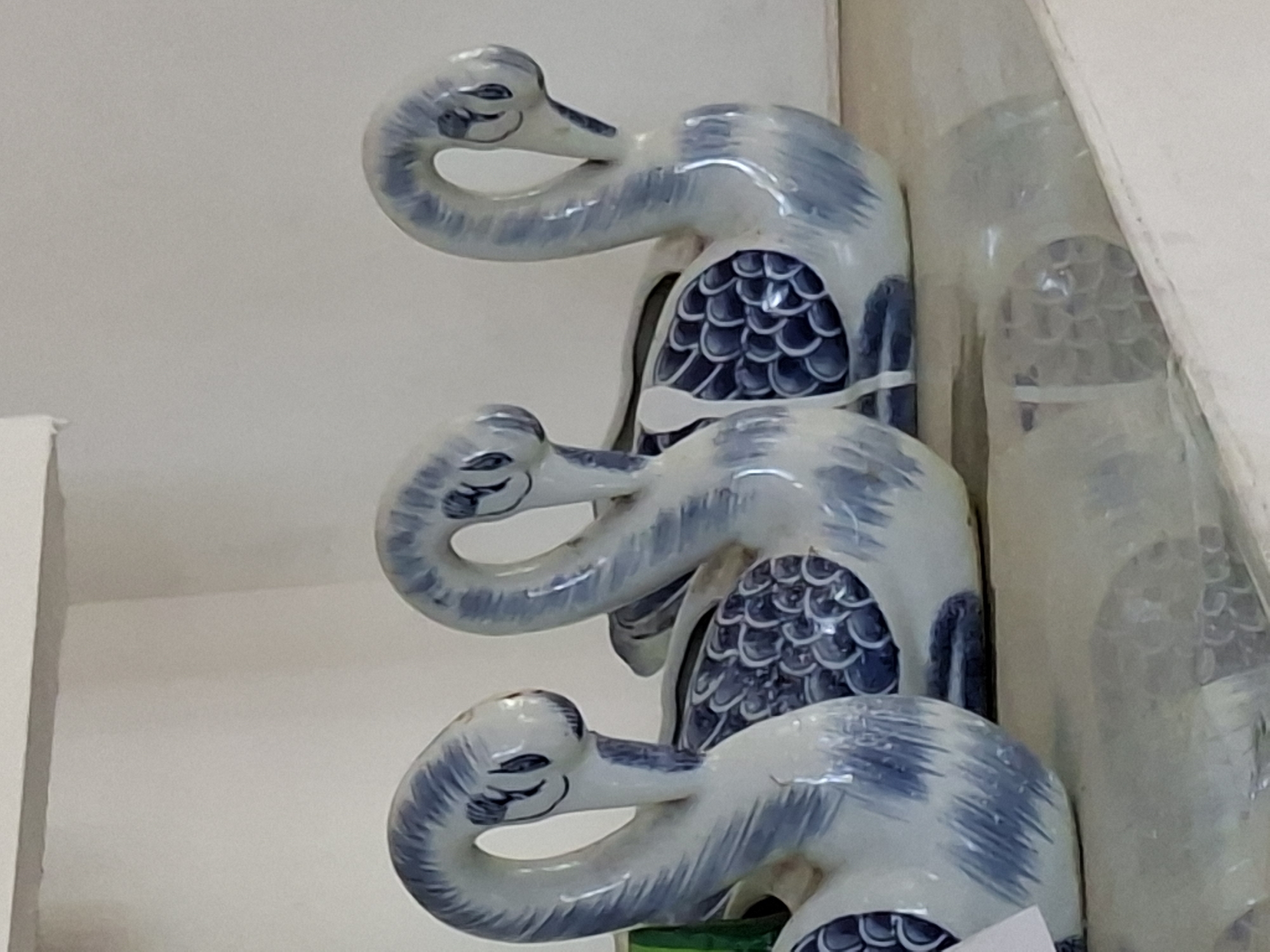 Three blue and white ceramic crane figurines are stacked vertically. The figurines are detailed with scales and have elongated necks that curve to form a hook-like head. They appear to be indoors. There is no indication of the city of Uleereuleung, Indonesia.