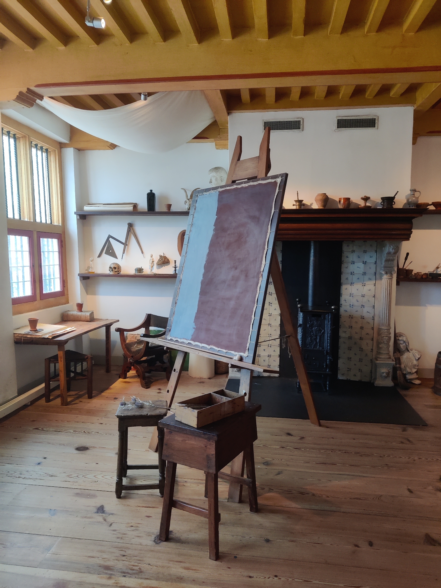 This image captures an artist's studio setting, likely a historical reproduction or a preserved space, within the city of Amsterdam, Netherlands. The room features a wooden floor with visible grain and some wear. A large easel stands prominently in the center, displaying a canvas that is partially covered with a reddish-brown and light blue paint.
To the left, a wooden desk with a simple stool is positioned near a window with multiple panes and red trim. On the desk, there are a few items, including a book and some papers. A wooden chair with an ornate design is situated behind the desk.
The back wall of the room is dominated by a fireplace, which has decorative tiles and a black stove. Above the fireplace, a wooden mantelpiece holds various pottery and decorative items. Shelves above the mantelpiece display more objects, including a rolled-up scroll, a black bottle, and what appears to be anatomical models or artistic references like a skull and a compass.
The ceiling is made of exposed wooden beams, with some fabric draped between them. Lighting fixtures are visible on the beams. The overall impression is one of a well-lit workspace, perhaps preserved to evoke a sense of historical artistic practice. There are no people visible in the image.