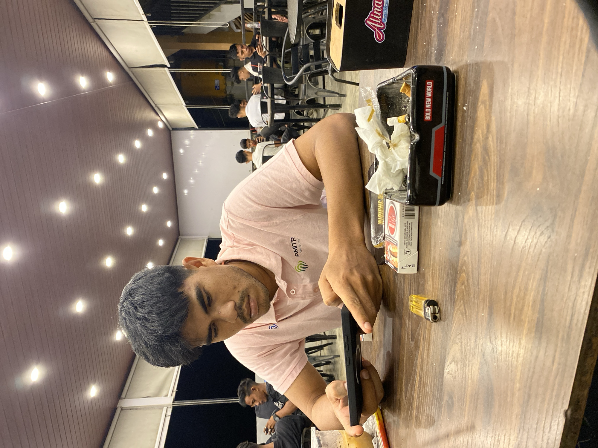 The image captures a casual evening scene at a cafe or restaurant in Keudeublang, Indonesia. The primary subject is a young man with dark hair and a light beard, wearing a pink polo shirt with "AMITR" and "syariah" visible on it. He is engrossed in his black smartphone, held in both hands, while seated at a wooden table.
On the table, an ashtray is prominently displayed, filled with cigarette butts and crumpled tissues, bearing the text "BOLD NEW WORLD." Beside it are a yellow cigarette lighter and a white cigarette pack showing the "BAT" logo. A black container with "Alinaa" written on it is also visible on the table.
In the background, several other young men are present, sitting at dark tables and chairs, engaging in conversation or waiting, suggesting a social gathering. The setting features a ceiling with multiple bright, recessed lights, indicating that the picture was taken at night or in the evening. The overall ambiance is relaxed and social.