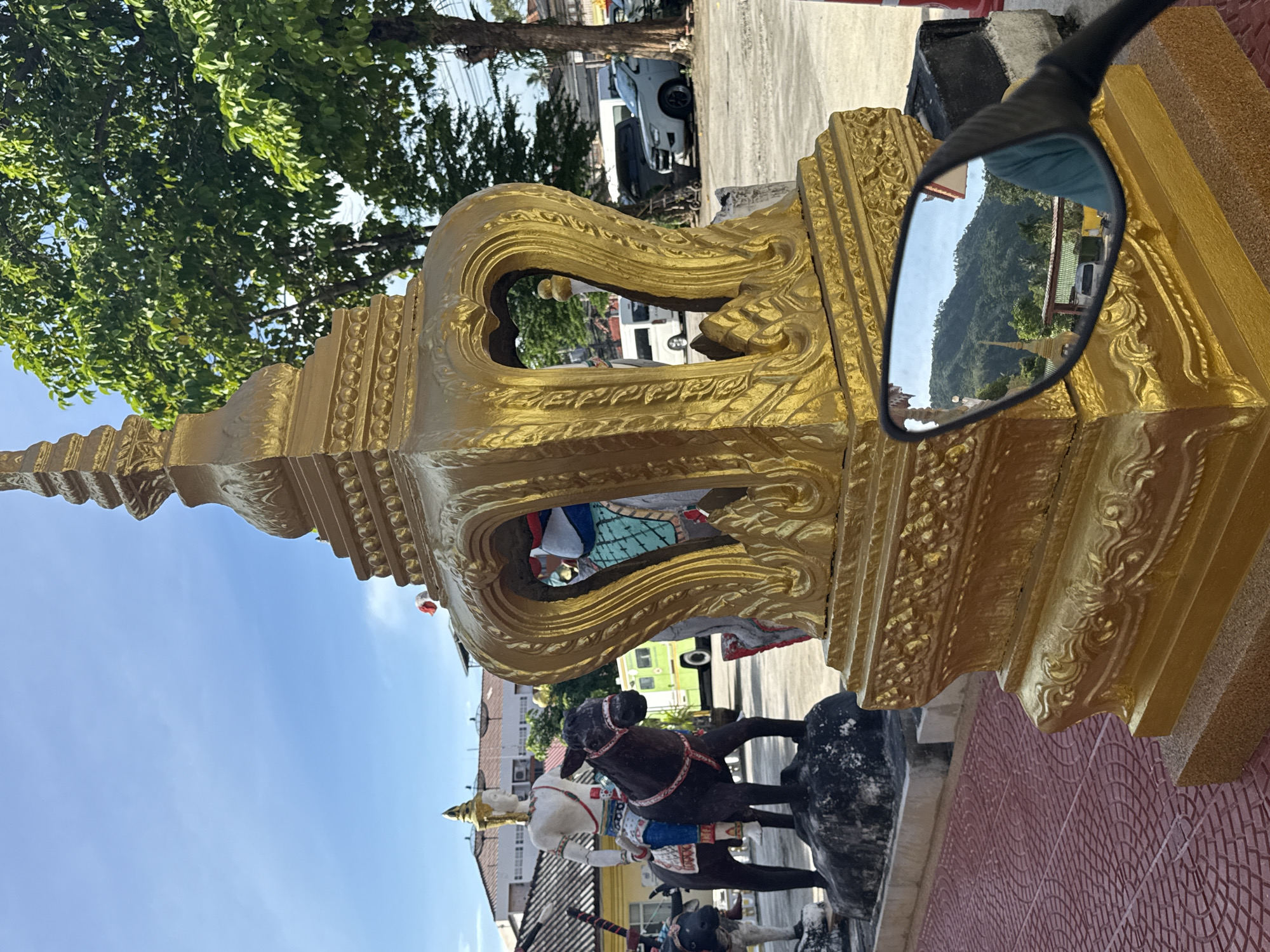 In Baan Hua Thanon, Thailand, a close-up shot focuses on a golden, ornate structure with intricate carvings, possibly part of a Buddhist temple or shrine. The structure is in the foreground, and a motorcycle's side mirror is attached to its base, reflecting a landscape of lush green hills and a golden stupa. In the background, a statue of a person riding a horse stands next to another statue of a bull. Other elements visible include trees, buildings, parked vehicles, and a blue sky with scattered clouds. The ground is paved with red tiles. The scene suggests a cultural and religious site, likely a tourist destination.