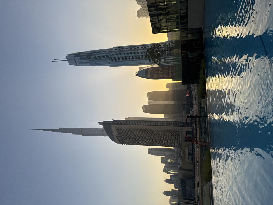 The image displays a panoramic view of the Downtown Dubai skyline in the United Arab Emirates, observed from an elevated perspective, likely a rooftop pool deck. The scene is bathed in the warm, golden light of either sunrise or sunset, creating a gradient from a pale blue sky at the top to a bright amber glow along the horizon.
In the foreground, a tranquil, blue-tiled infinity-edge swimming pool occupies the lower half of the frame. Its surface reflects the surrounding city and sky, exhibiting minor ripples. Partially submerged on the left side of the pool is a red-orange pool ladder or access steps. Beyond the pool's edge, a narrow band of dark, low-lying vegetation is visible, separating the pool area from the midground.
The midground features a multi-lane road with visible vehicular traffic, running parallel to a cluster of multi-story commercial or residential buildings. One prominent building in this section displays an "EMAAR" logo.
The background is dominated by the dense cluster of high-rise skyscrapers forming the Dubai skyline, largely silhouetted against the bright horizon. The most prominent structure, the Burj Khalifa, is situated near the center-left with its distinctive spire extending into the pale blue sky. To the right of the Burj Khalifa, another tall, slender skyscraper, identified by an "EMAAR" logo near its peak, stands out. Additional skyscrapers of various designs and heights are visible across the horizon, some appearing dark and reflective. No individuals or specific actions are discernible within the frame.