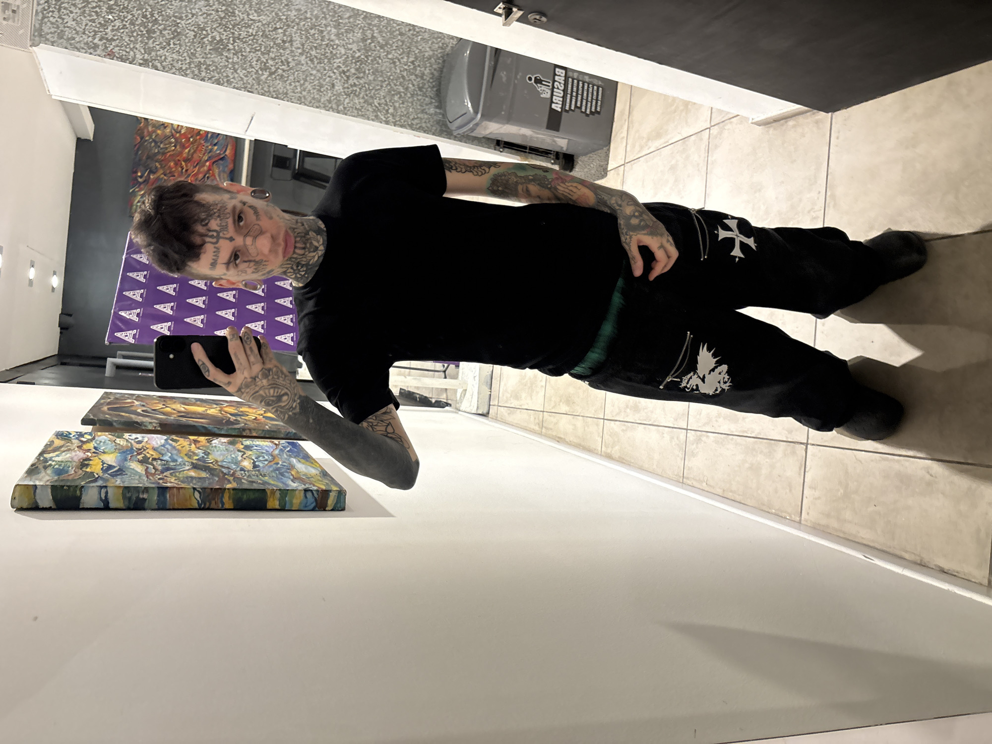 A man with numerous face and body tattoos stands in an art gallery, holding a smartphone, presumably taking a selfie. He is wearing a black t-shirt and black pants with white cross designs. Behind him are several paintings on display. A grey trash receptacle is located further back in the scene. The location may be in Buenos Aires, Argentina.