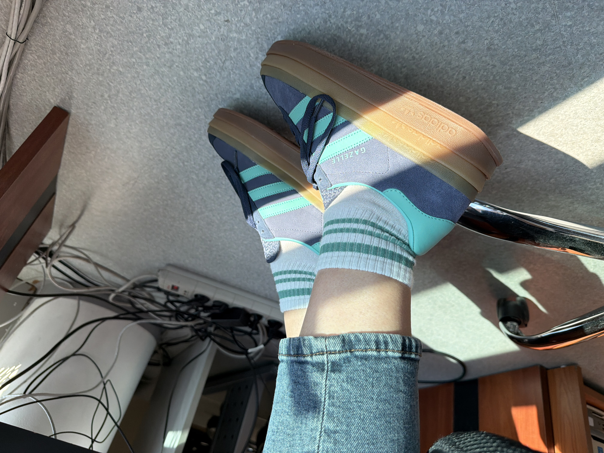 This image depicts a close-up view of a person's feet wearing blue denim jeans and Adidas Gazelle platform sneakers with mint green accents. The person is seated in an office chair with a chrome base. The background shows an office environment with wooden furniture, a white cylindrical object with black wires, and a power strip. The floor appears to be linoleum. There is sunlight casting shadows on the floor and the chair. The scene is indoors, in what appears to be an office setting. There is no specific indication of the city of Sofia, Bulgaria.