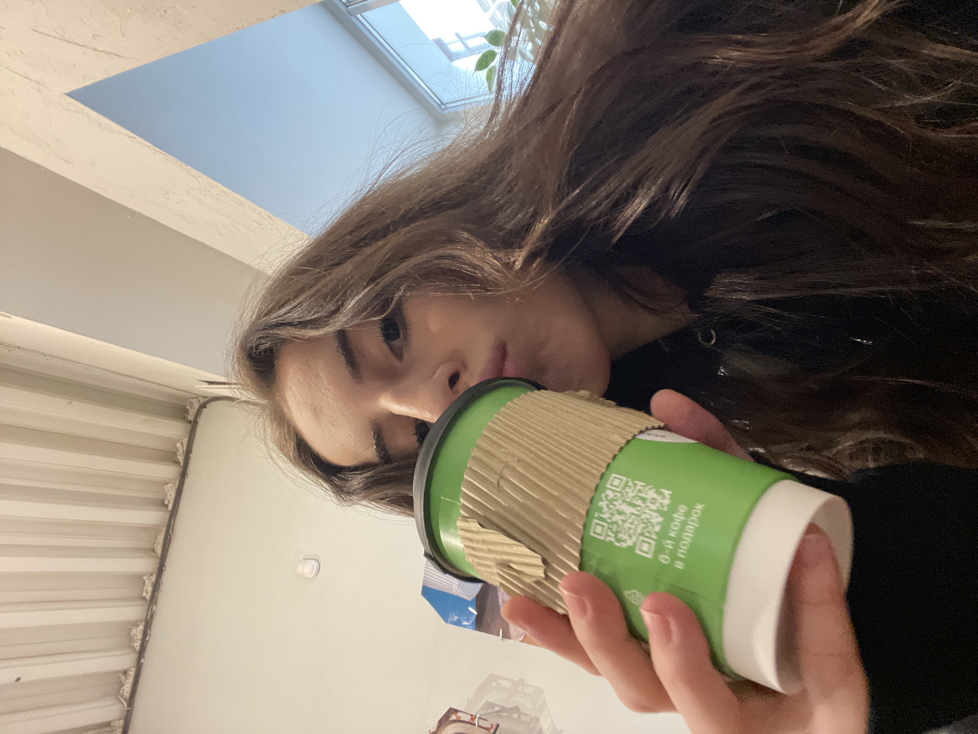 A young woman is holding a green and white coffee cup, with a brown cardboard sleeve. The cup has a QR code and text in Russian that reads "0-й кофе в подарок" (0th coffee as a gift). The woman has long brown hair, and she is wearing a black jacket. The background appears to be the interior of a building in Moscow, Russia. There is a window with a view of the sky.