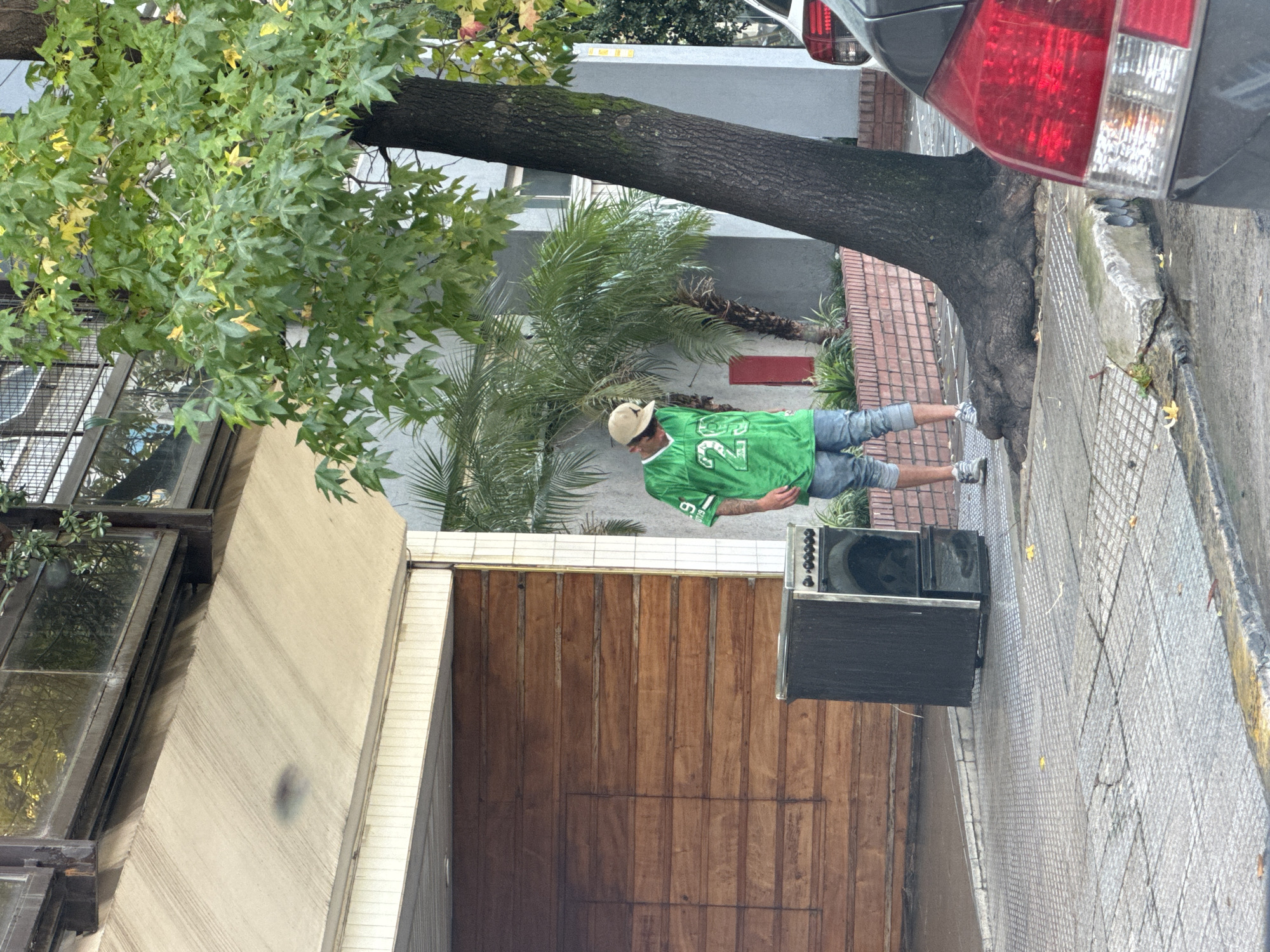 A street scene in Buenos Aires, Argentina, shows a person wearing a green sports jersey, shorts, and a hat standing on a sidewalk next to a large tree and a dark-colored speaker. The person is positioned near a wooden garage door and tiled sidewalk. The foreground includes a light-colored building and foliage. A car is parked on the street. The scene appears to be taken during the day in cloudy conditions.