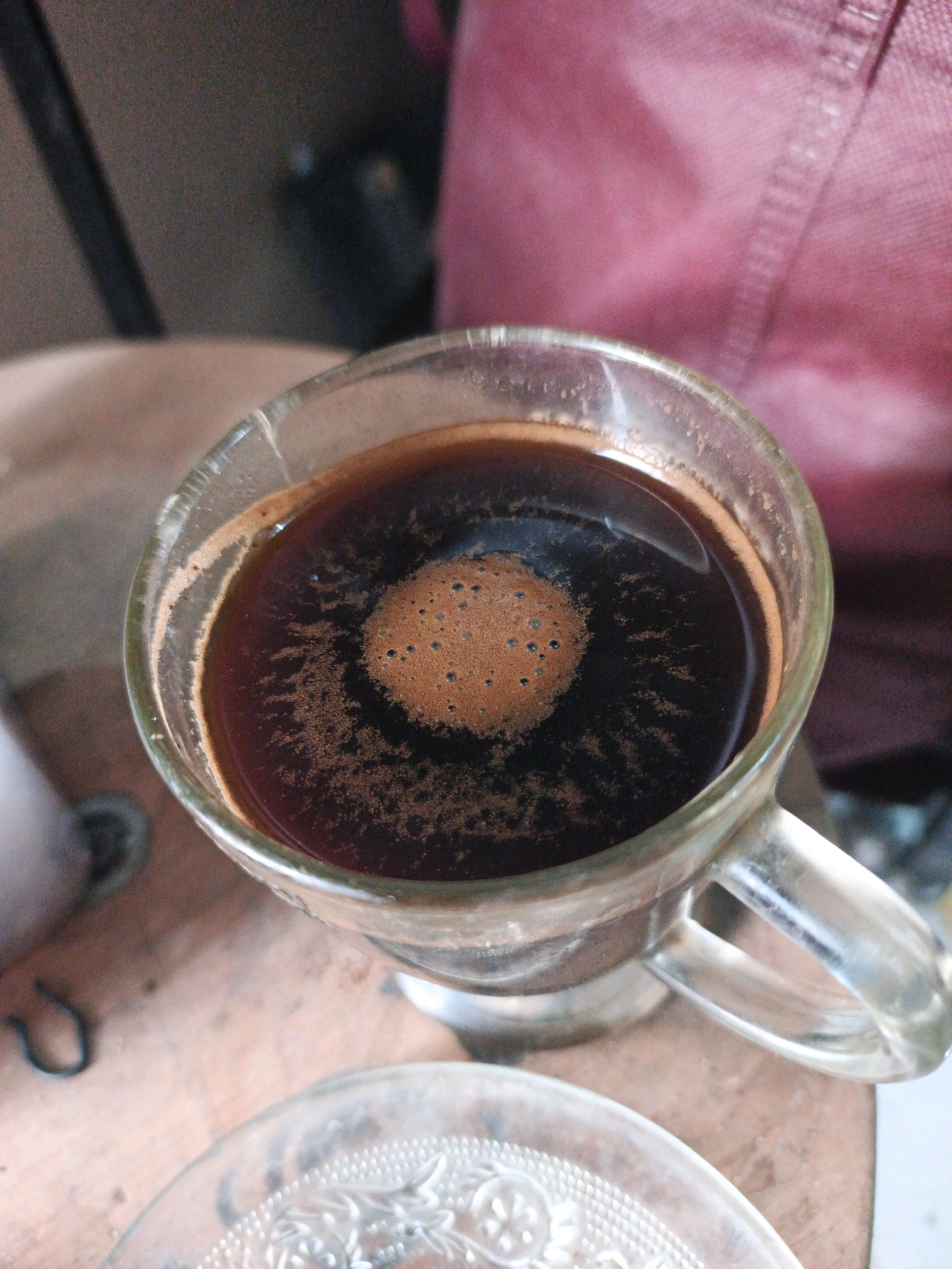 A close-up, overhead shot captures a glass mug filled with dark coffee, topped with a frothy, light brown crema. The crema forms a circular pattern in the center of the mug, with a textured, bubbly surface. The mug has a clear glass handle and sits on a light brown, wooden surface. In the background, out of focus, is a textured pink fabric. There is also a glimpse of a clear glass saucer with an embossed floral pattern in the lower foreground. The scene appears to be in Ketegan, Indonesia, with the visual cues suggesting a domestic or casual setting. No individuals are visible in the image.
