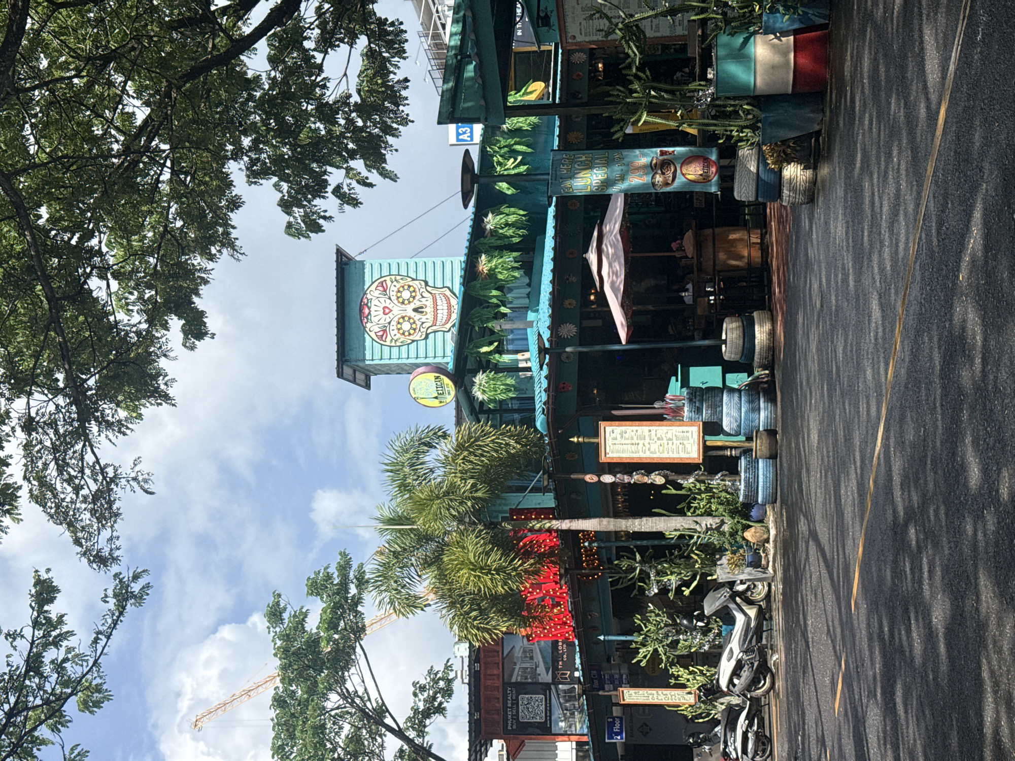 The image depicts a vibrant street scene in Choeng Thale, Thailand, featuring a colorful building with a large sugar skull decoration on its facade. The building appears to be a restaurant or bar, with outdoor seating, umbrellas, and a menu displayed. In front of the building, there are several motorcycles parked along the roadside, as well as potted plants, including cacti and palm trees. The street in the foreground is paved and wet, reflecting the dappled sunlight filtering through the lush green trees overhead. A construction crane is visible in the background, suggesting ongoing development in the area. The overall atmosphere is lively and eclectic, with a blend of architectural styles and cultural influences.