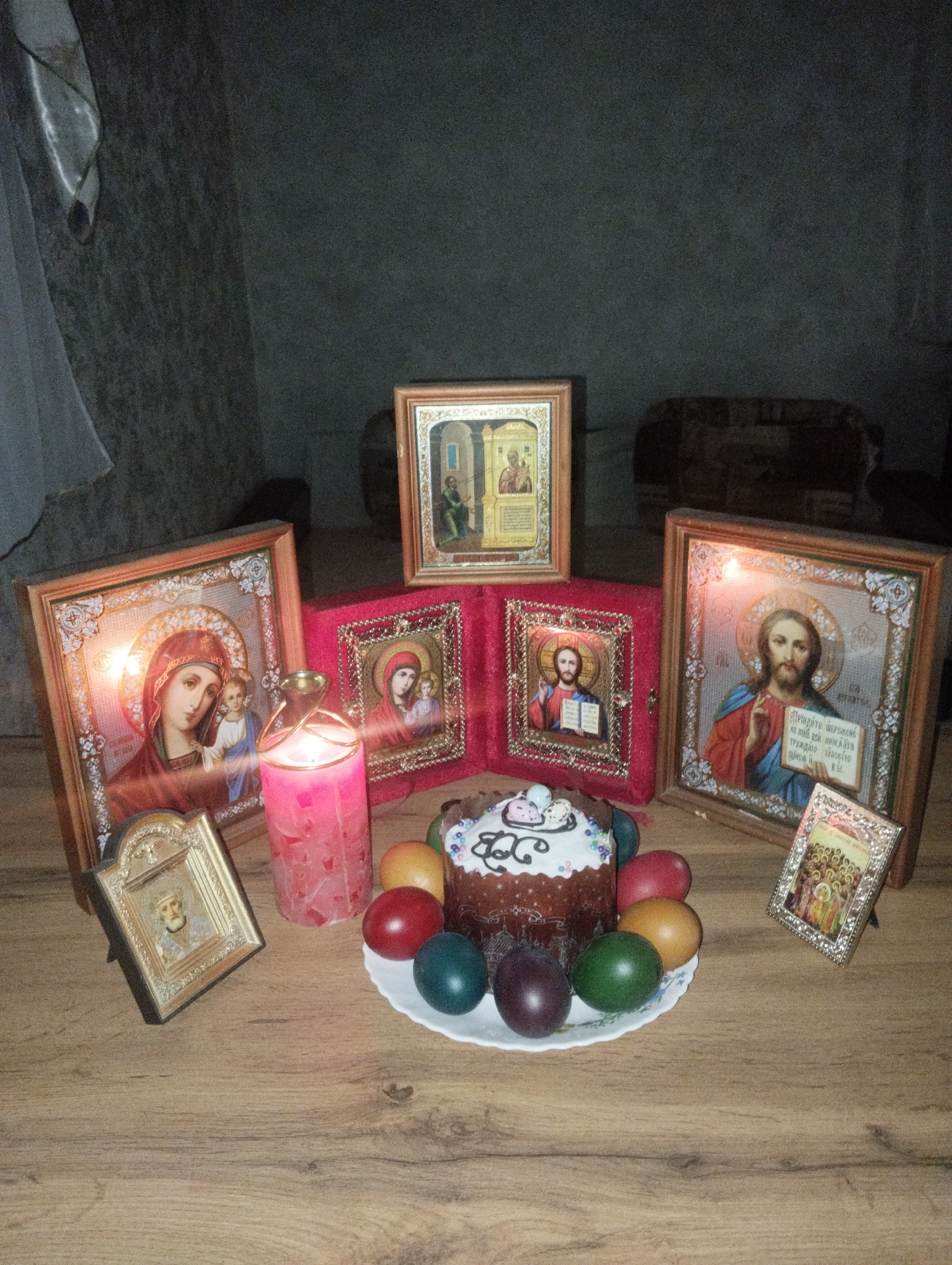 A table is set with religious iconography, a pink candle, an Easter cake topped with white icing and decorative elements, and colored eggs. The framed religious icons are arranged around the cake, and the candle provides a central light source. The eggs are painted in shades of green, red, gold, and maroon. The location is Grishanki, Russia.