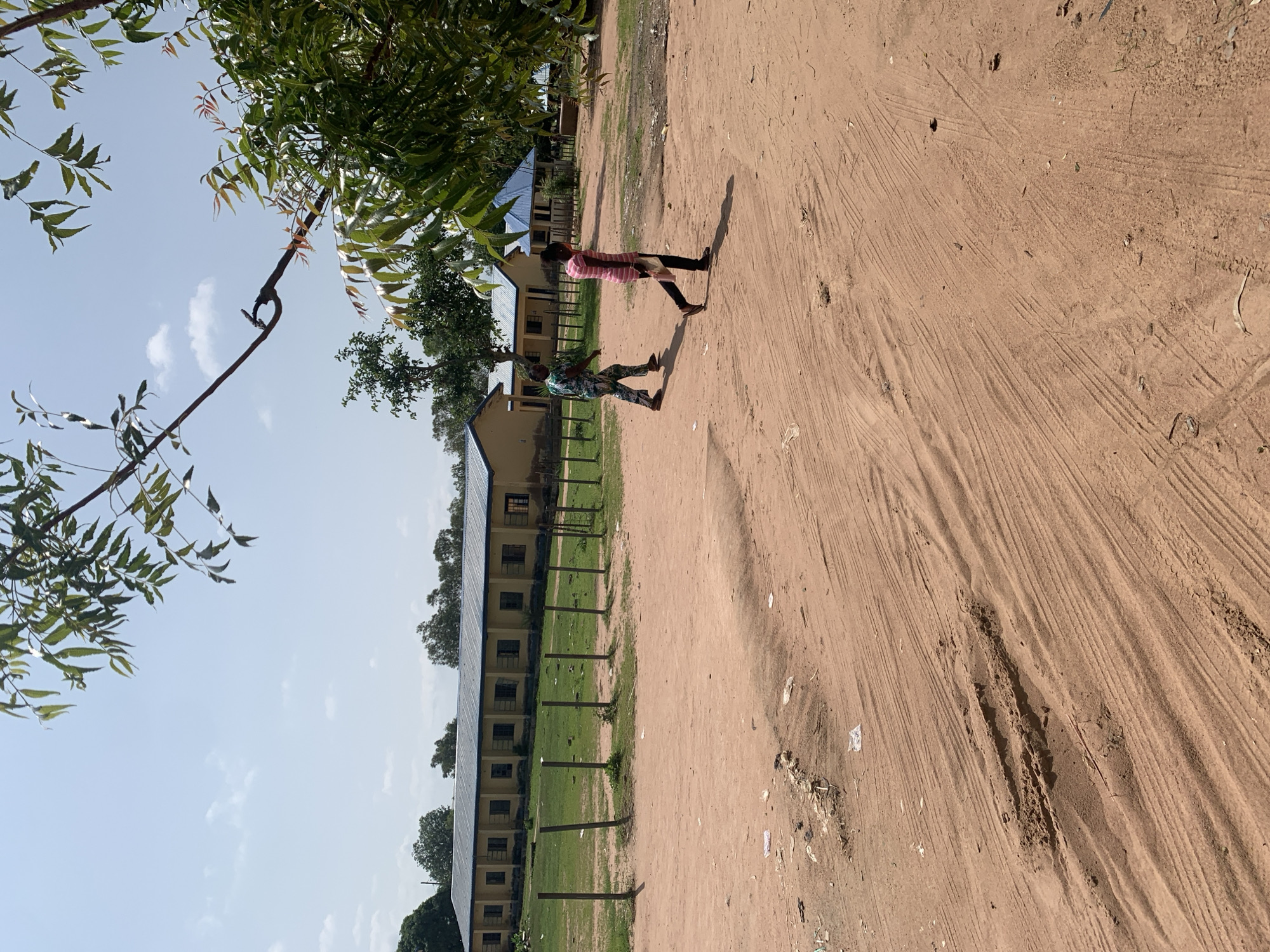 Two individuals are walking on a dirt road in front of a yellow building with a blue roof. The building appears to be a school or administrative facility with multiple windows. The ground is sandy with tire tracks and some litter. Trees are visible in the background. The sky is blue with a few clouds. The scene is likely located in Jalingo, Nigeria. The person on the left is wearing a multicolored patterned outfit, and the person on the right is wearing a pink and white striped shirt and black pants.