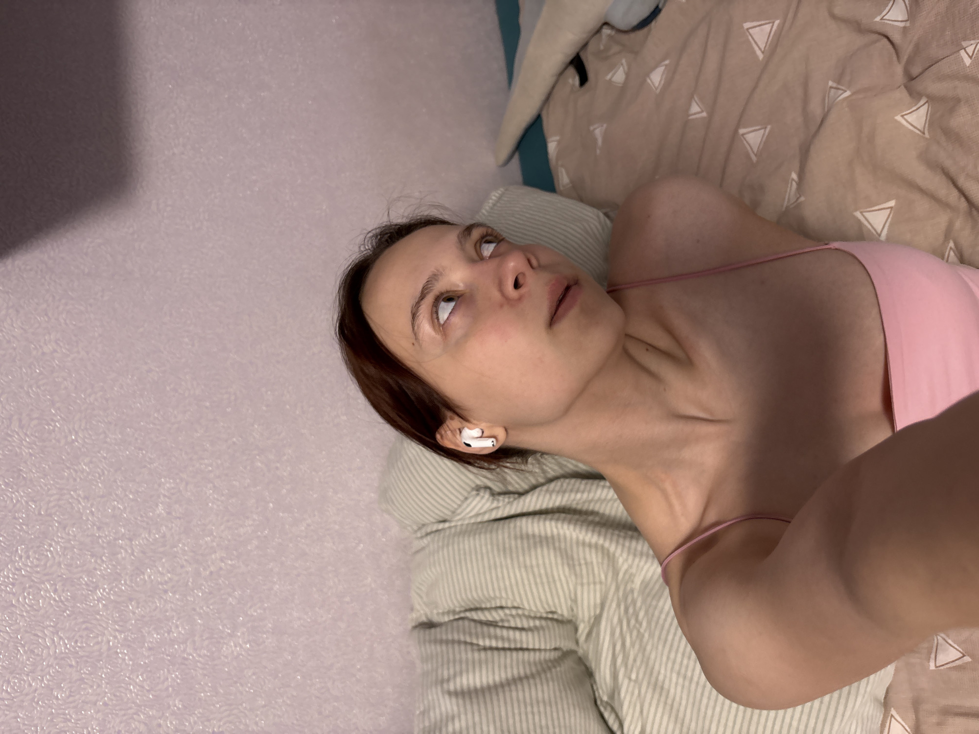 A woman with brown hair and fair skin is lying on a bed. She is wearing a pink top and has a white earbud in her right ear. She is looking upwards and to the left. The bed has a striped pillow and a brown comforter with white triangles. The wall to the left of the bed is textured and light purple. The location is likely indoors, possibly in a residential area in Прикамский, Russia, based on the user's location.