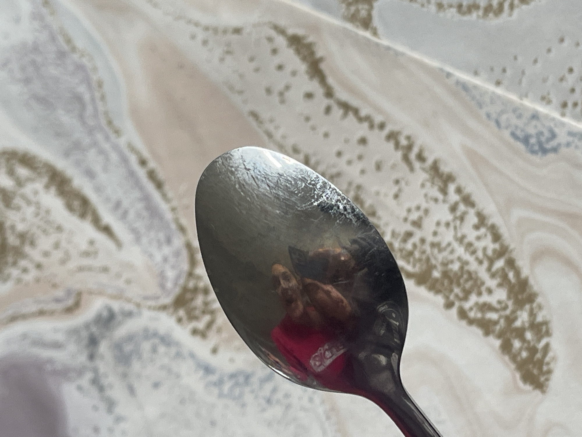 A close-up shot depicts a shiny metal spoon reflecting a person wearing a pink shirt and holding a phone. The background appears to be a surface with a marbled pattern in shades of white, beige, and gray. There is no information to suggest this is in Saarbrücken, Germany.