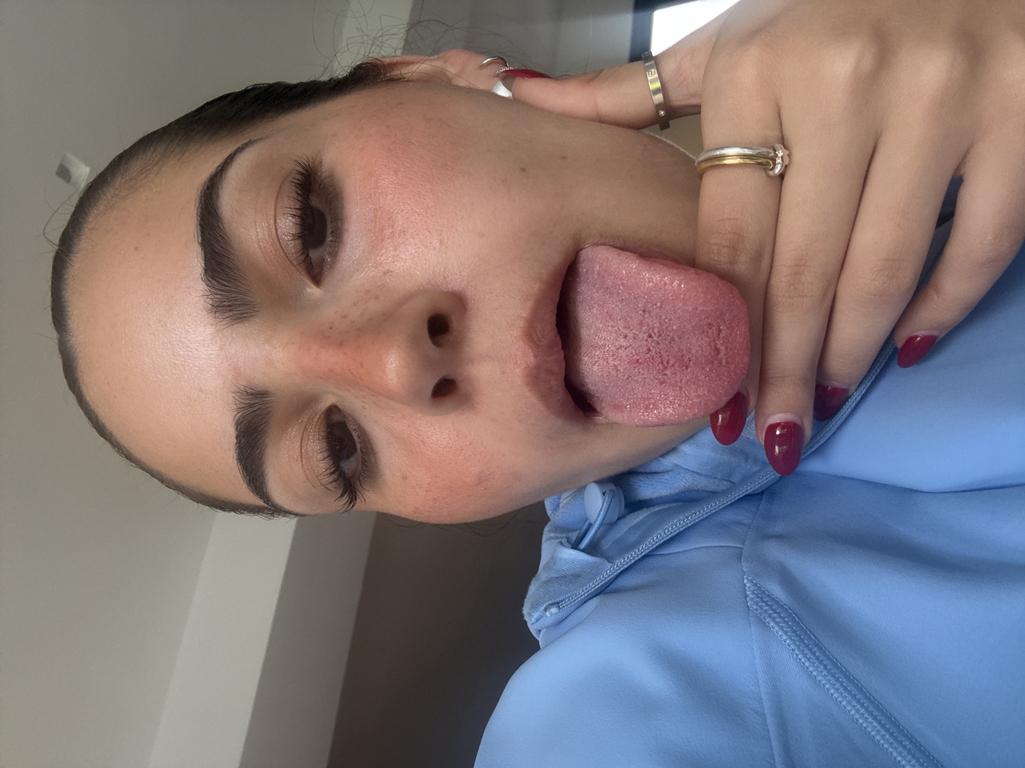 A close-up, eye-level, selfie-style shot shows a young woman indoors, sticking her tongue out. She is wearing a light blue hooded jacket and has bright red nail polish. She has defined eyebrows, dark eyelashes, and is wearing subtle makeup with rosy blush on her cheeks. She is also wearing several rings on her fingers, including a thin band with a circular emblem and a two-toned band. The background appears to be a room with a plain ceiling. There is no visible indication of San Bernardino Tlaxcalancingo, Mexico.