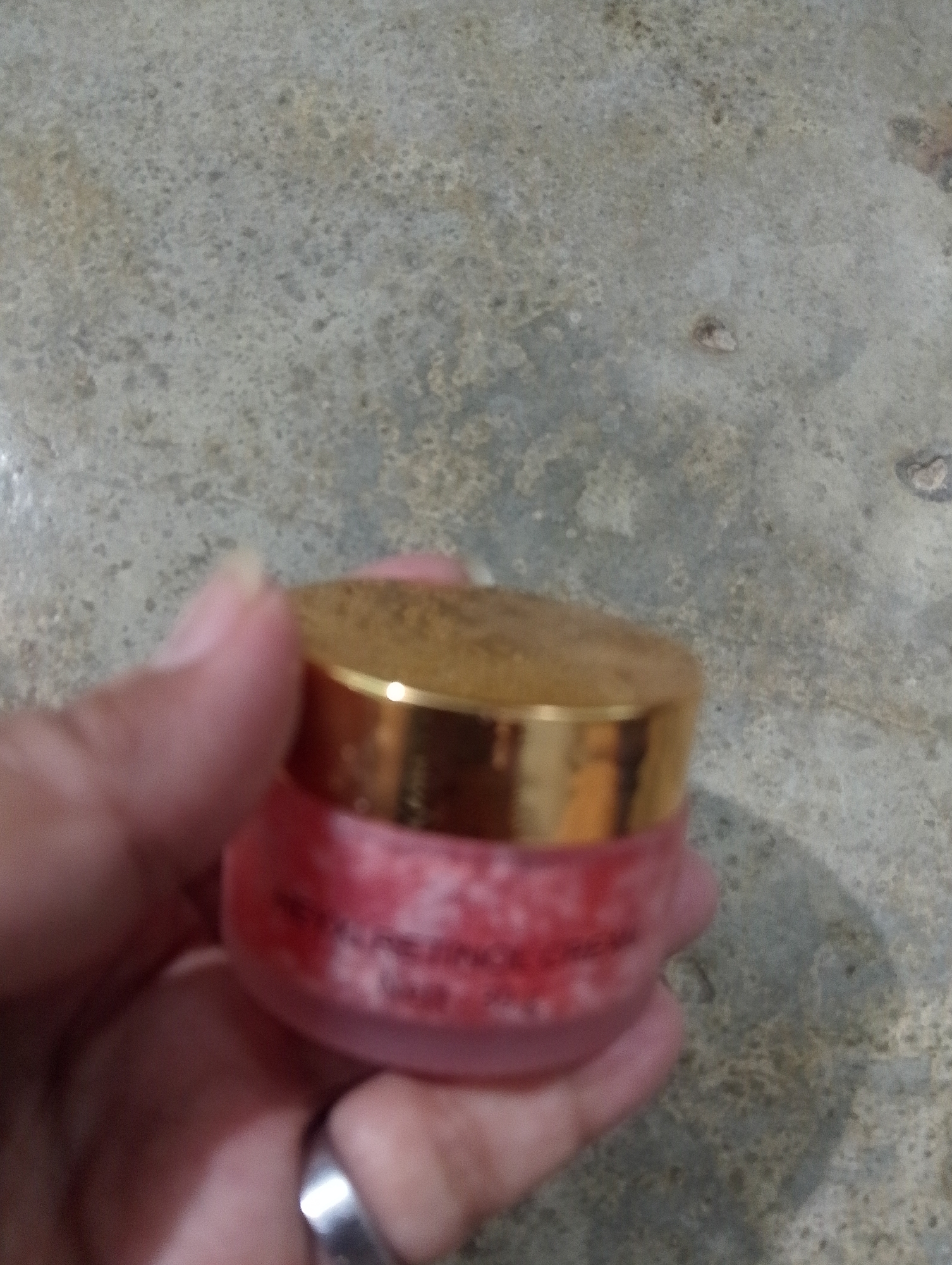 A close-up shot shows a hand holding a small, round cosmetic container with a gold lid. The container appears to be filled with a pink substance that has a textured, granular appearance. The background is a textured gray surface, possibly concrete or stone. The image is slightly out of focus, particularly the background. There are no specific landmarks or contextual clues within the image to indicate the city of Serbelawan, Indonesia.