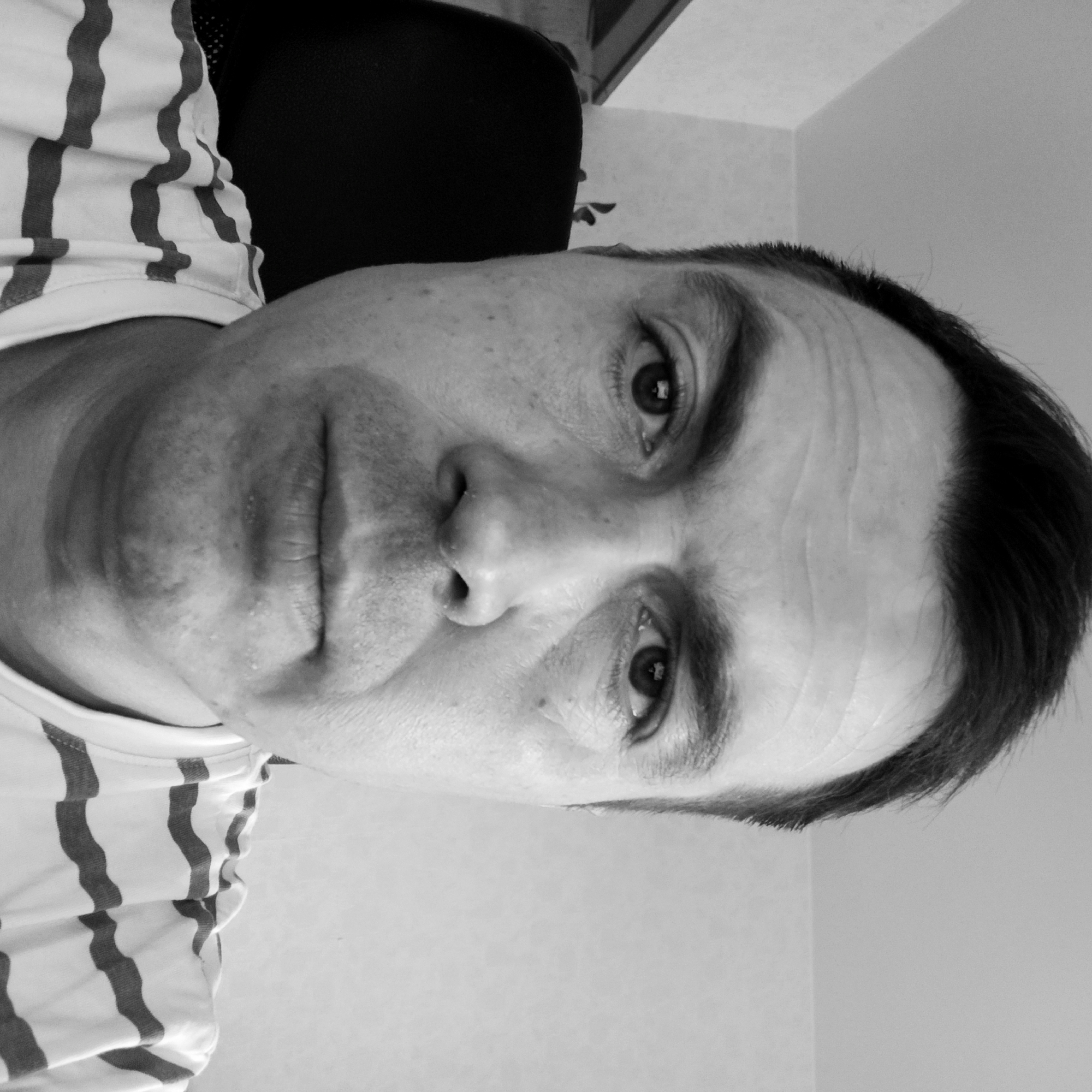 A black and white, close-up, head-and-shoulders selfie of a man with dark hair. He is looking directly at the camera. He is wearing a striped t-shirt. He is seated in a chair in what appears to be an indoor setting, with a wall visible behind him. The background is out of focus. There is no discernible location in Nizhny Novgorod, Russia.