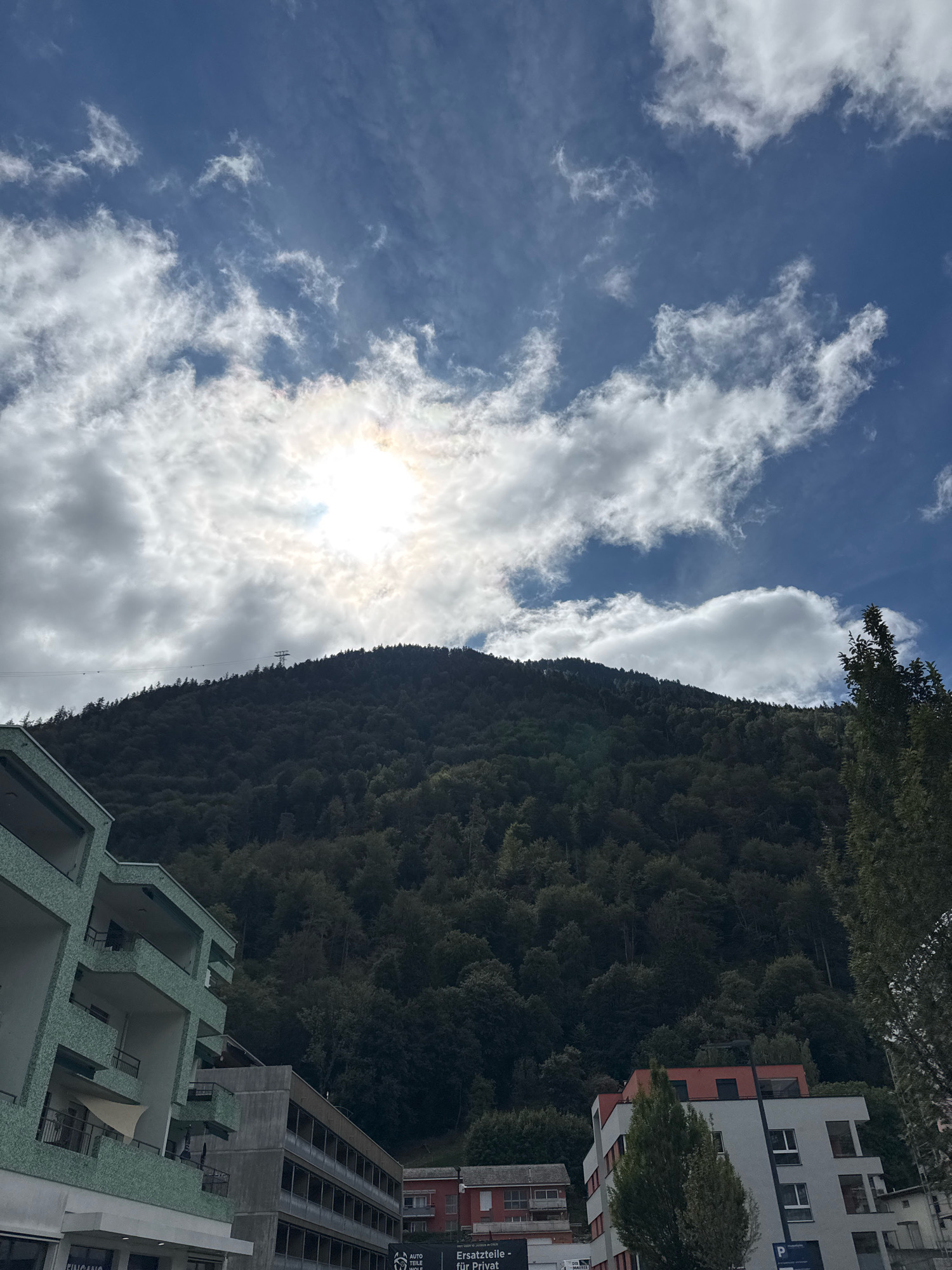 The image shows a mountain covered in dense green forest, reaching towards a sky with a mix of bright sun and scattered white clouds. In the foreground, there are several buildings, including a modern apartment building with a light green facade and balconies, a multi-story concrete building, and other smaller structures. A cable car line with towers and cables is visible stretching across the side of the mountain. The scene appears to be an urban or semi-urban area at the base of a mountain.