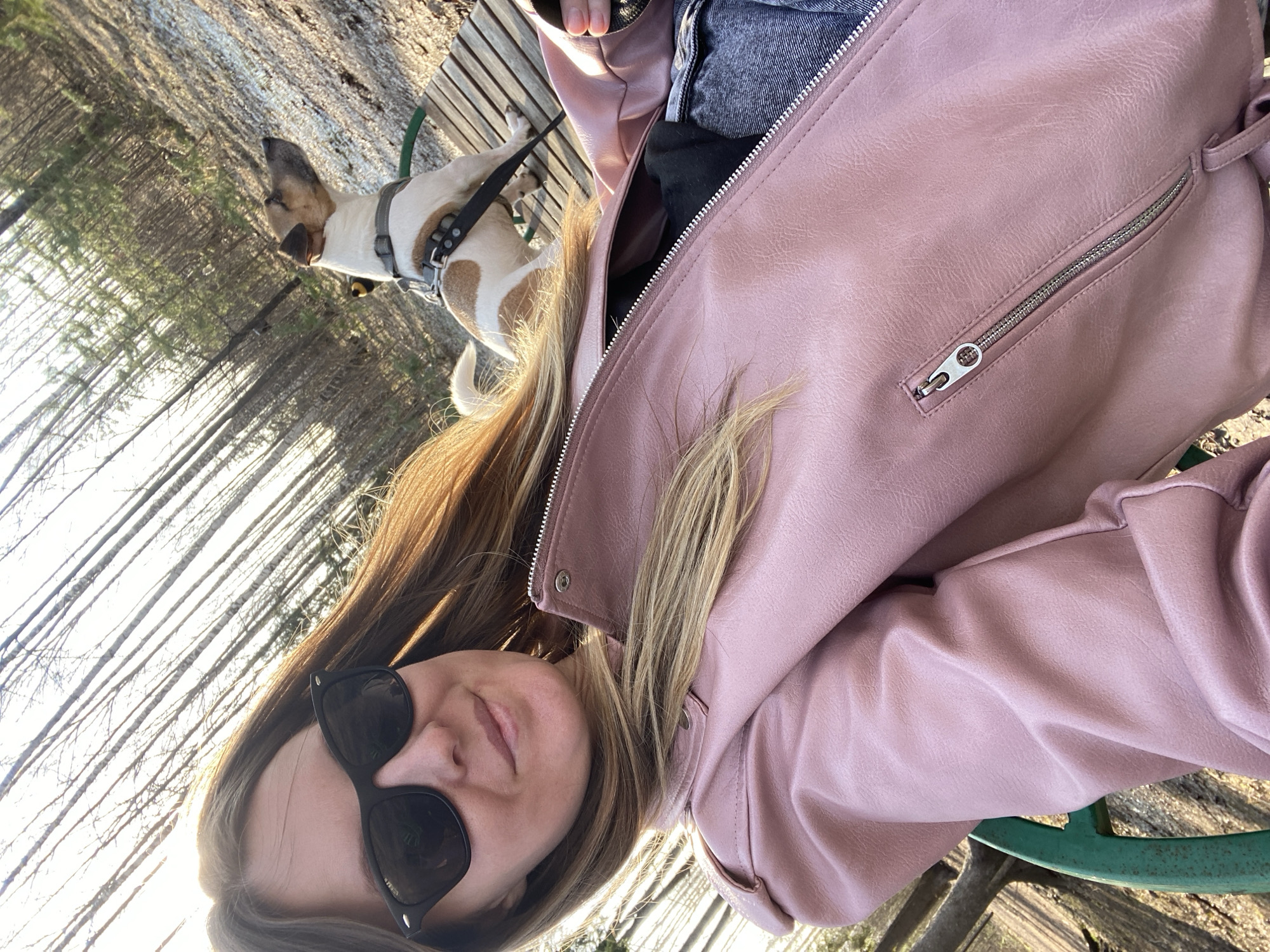 The image shows a woman seated on a park bench next to her dog. She is wearing sunglasses and a pink jacket. Her dog is small, white, and tan and is leashed. The location is a wooded park with tall, bare trees, typical of a landscape near Noviy, Russia.