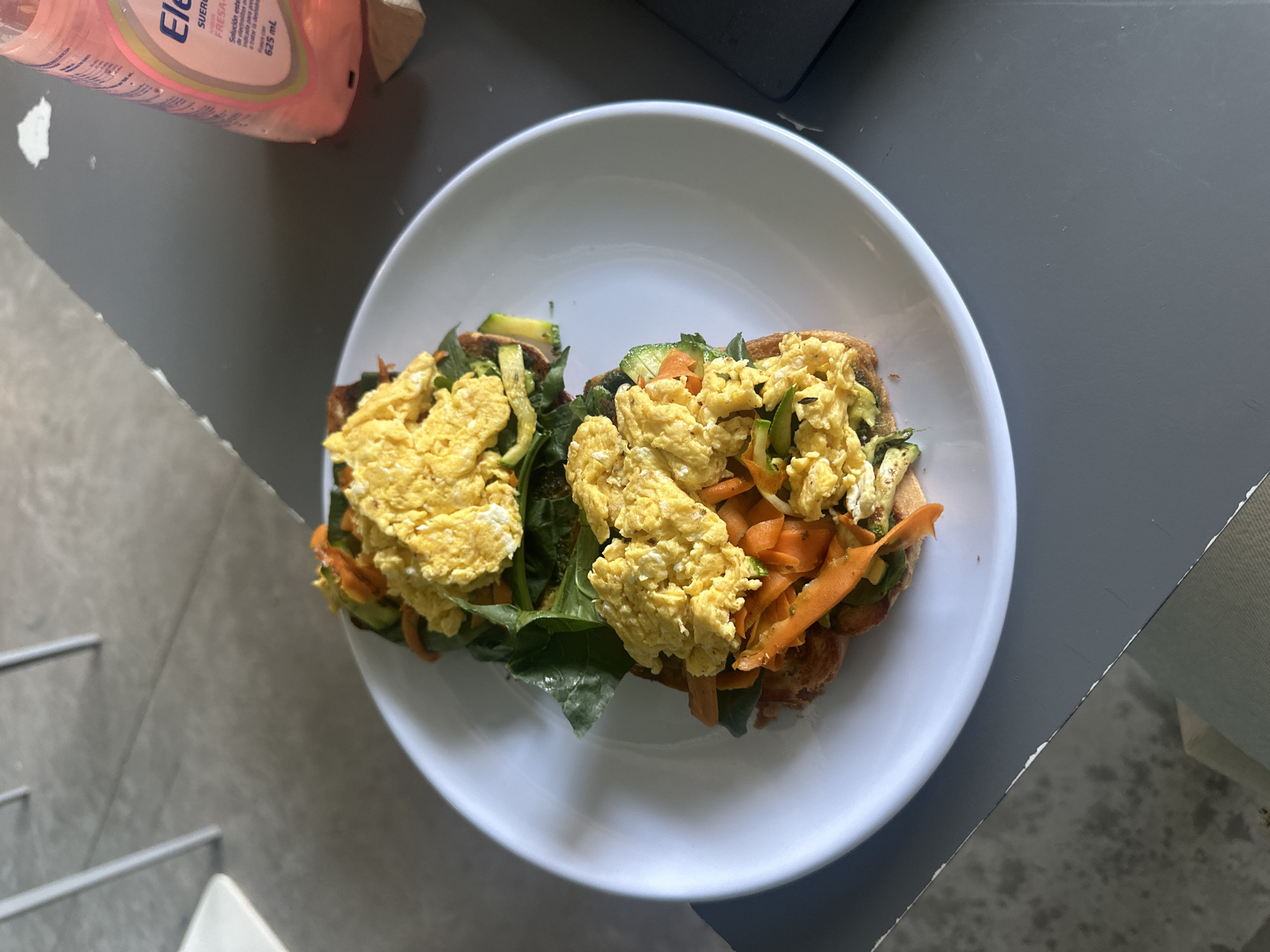 The image shows a white plate with two pieces of toast topped with green leafy vegetables, carrot strips, zucchini slices, and scrambled eggs. A pink bottle of electrolyte solution sits partially visible at the top left of the frame. The plate rests on a gray surface next to a white surface. The setting appears to be indoors, potentially in a dining area. There is no evidence to suggest that this image was taken in Mexico City, Mexico.