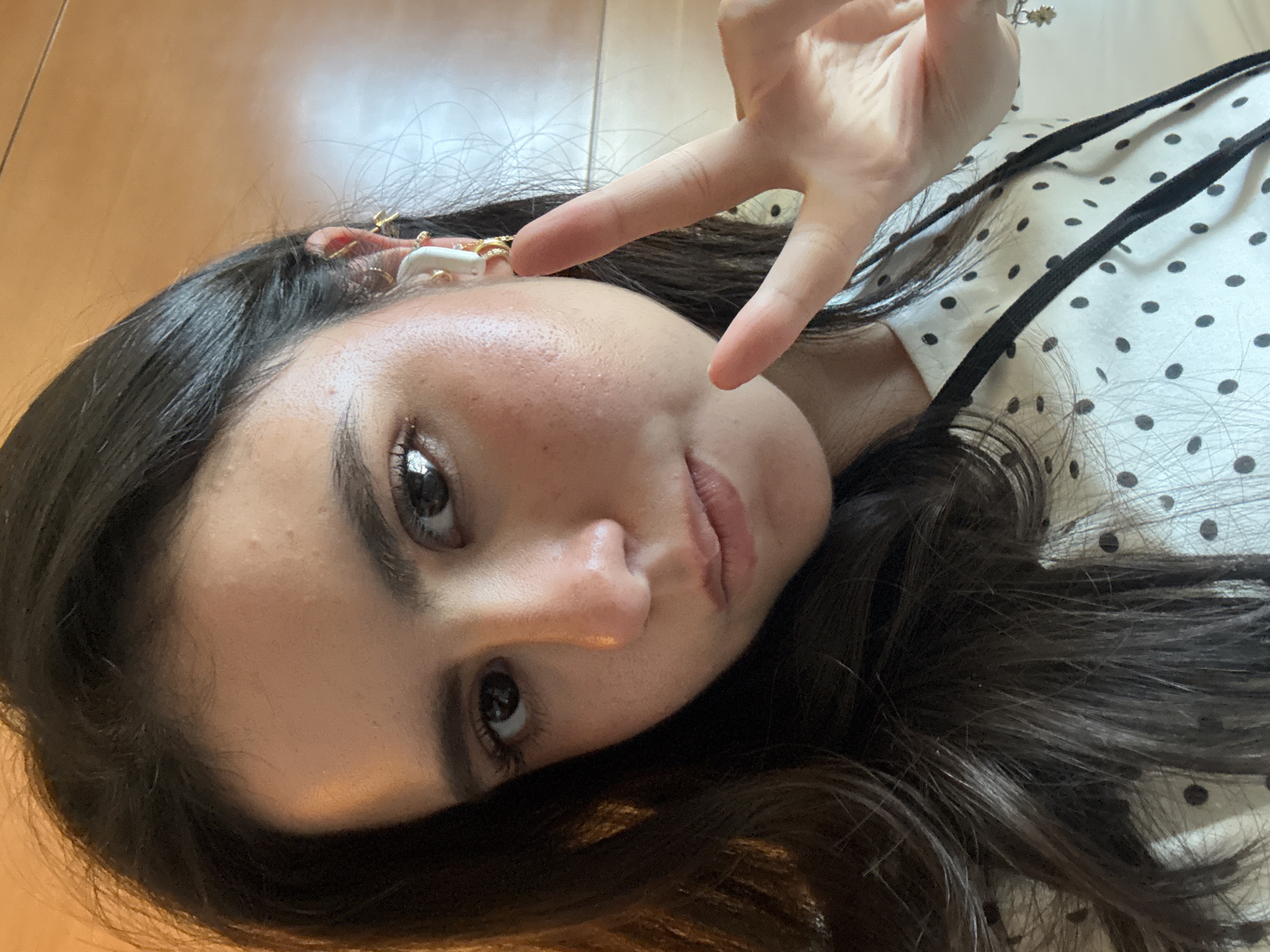 A young woman with long, dark hair takes a selfie in what appears to be a hotel room. She is wearing a white shirt with black polka dots and has on multiple earrings, including a white earbud in her right ear. Her right hand is raised, forming a peace sign, with her fingers partially obscuring her face. The background is blurred and appears to be a wall with warm lighting. There is no indication of the specific location in Los Angeles, United States of America, as the image does not contain any identifying landmarks or context.