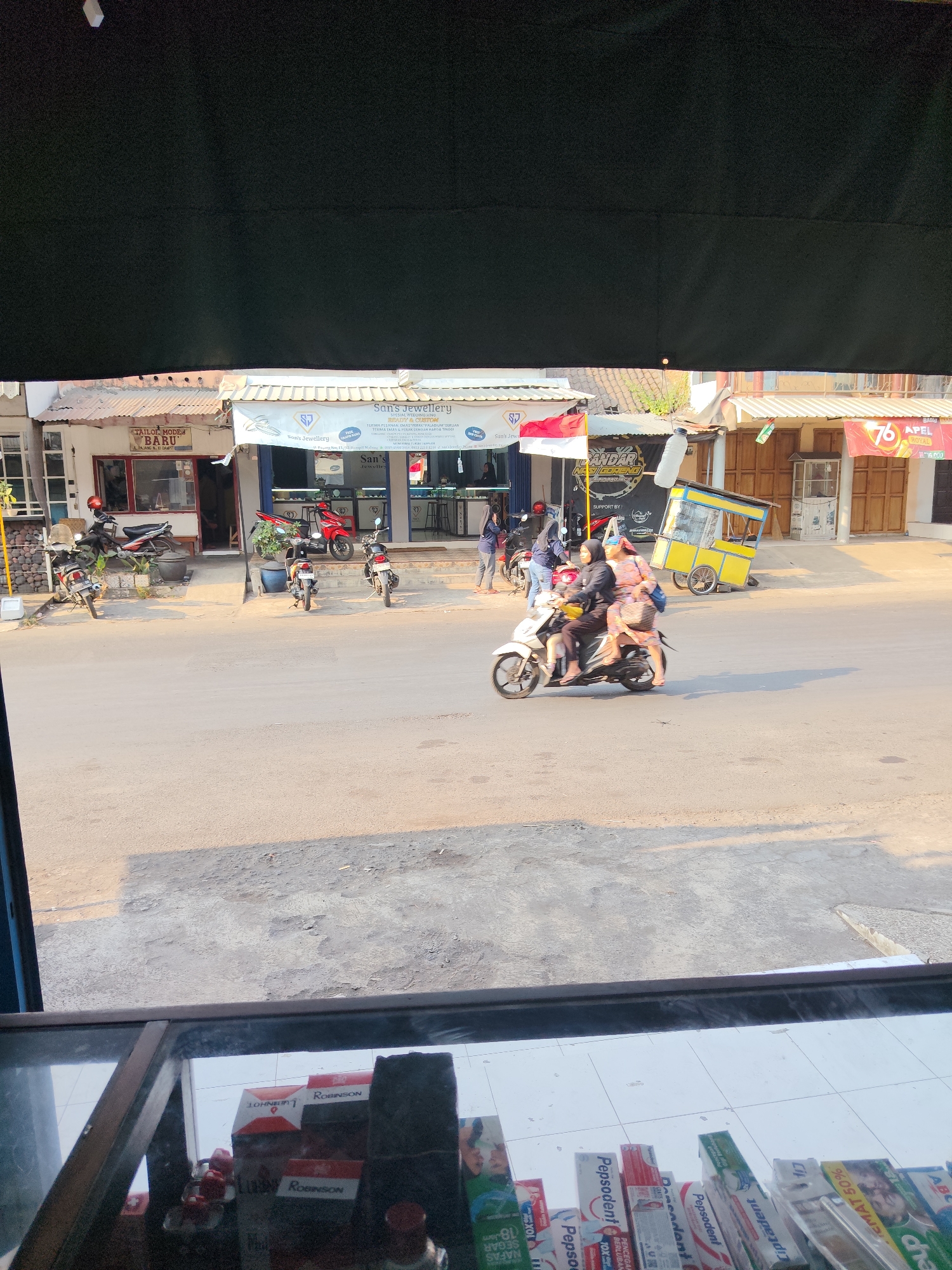 The image depicts a street scene in Dampit, Indonesia, in front of several shops. On the left, a shop labeled "TAILOR MODE BARU" is visible. Adjacent to it is "San's Jewellery" with a banner indicating "SPECIAL WEDDING RING". Further down the street, other businesses are present, including one with a sign reading "ANDAR NASI GORENG". A yellow food cart is parked on the sidewalk. Several motorcycles are parked along the storefronts and on the street. In the foreground, a woman and a child are riding a white motorcycle, and another motorcycle with two people is in motion. The street is paved, and the buildings have tin roofs and wooden doors. The lighting suggests it is daytime, with sunlight casting shadows. In the lower part of the image, a display case with various products, including toothpaste boxes, is visible, suggesting the perspective is from inside another shop looking outwards.