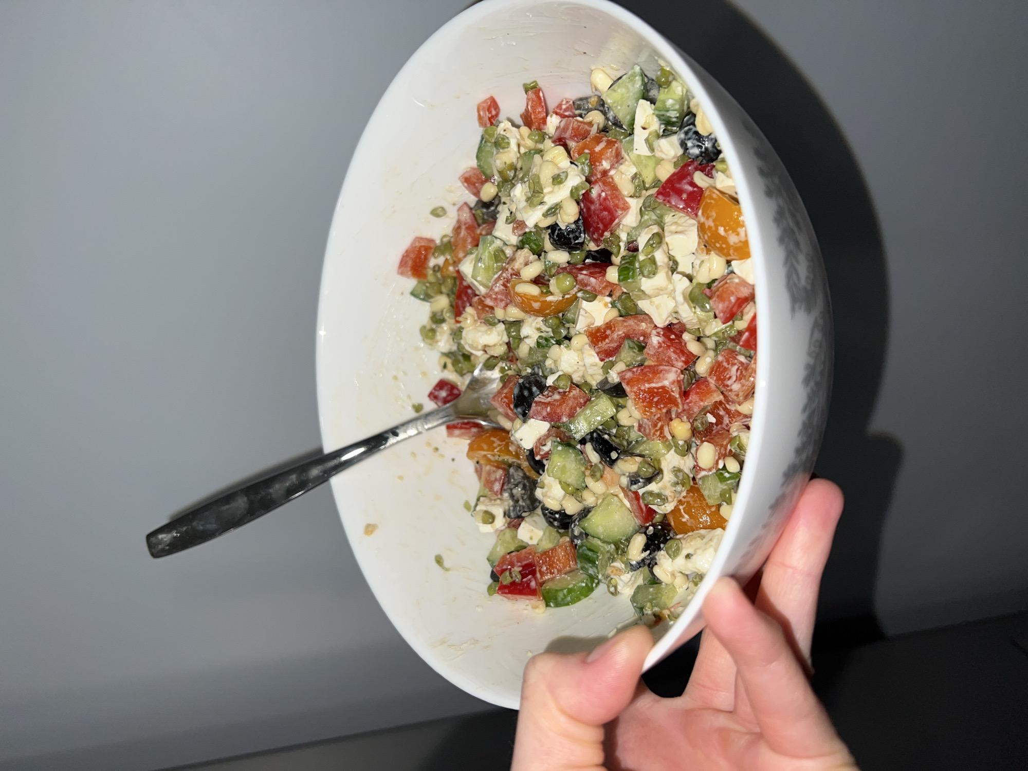A person's hand holds a white bowl filled with a salad. The salad consists of diced red and yellow tomatoes, cucumbers, feta cheese, black olives, and seeds, tossed in a dressing. A spoon rests inside the bowl. The background consists of a light gray wall with a dark gray surface below. There is no indication this image was taken in Khimki, Russia.