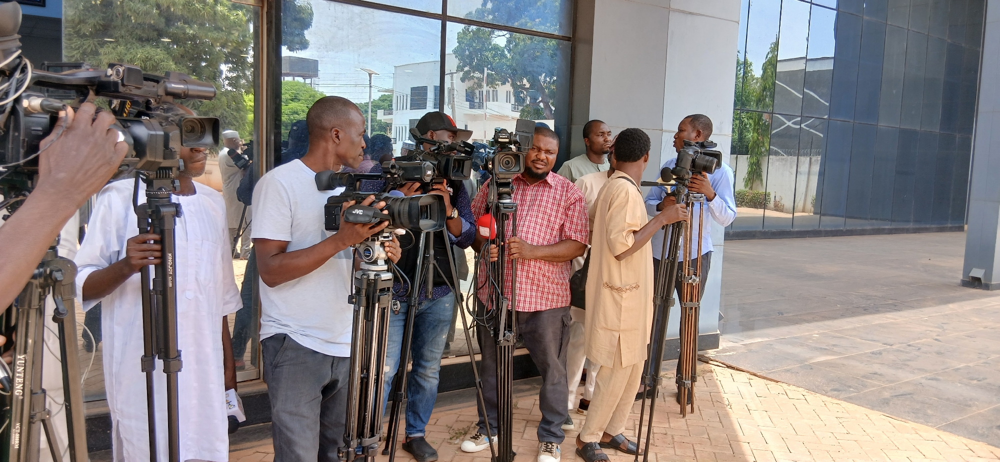 This media file captures a group of approximately eight to nine men, primarily camerapersons, gathered outdoors with their professional video equipment. The scene is set on a paved area in front of a modern building featuring large, reflective glass windows, suggesting an urban environment in Unguwar Shanu, Nigeria. The bright sunlight and clear reflections indicate it is daytime with good weather.
The men, mostly young to middle-aged, are dressed in a mix of casual attire, including t-shirts, jeans, and plaid shirts, as well as traditional Nigerian garments such as a white agbada and a light brown caftan/tunic. They are actively operating, setting up, or waiting with their cameras, suggesting they are covering an event, preparing for a press conference, or engaging in a video shoot. Their expressions appear focused and composed.
Numerous professional video cameras are visible on sturdy tripods. Recognizable brands include "Panasonic," "JVC," and "Sony." The tripods themselves bear brand names like "KING JOY" and "YUNTENG VC-888RM." One of the setups clearly shows a red microphone attached, indicating audio recording capabilities. The reflective glass facade of the building casts inverted reflections of surrounding trees and other structures, adding depth to the background. The ground is paved with light-colored tiles or bricks.