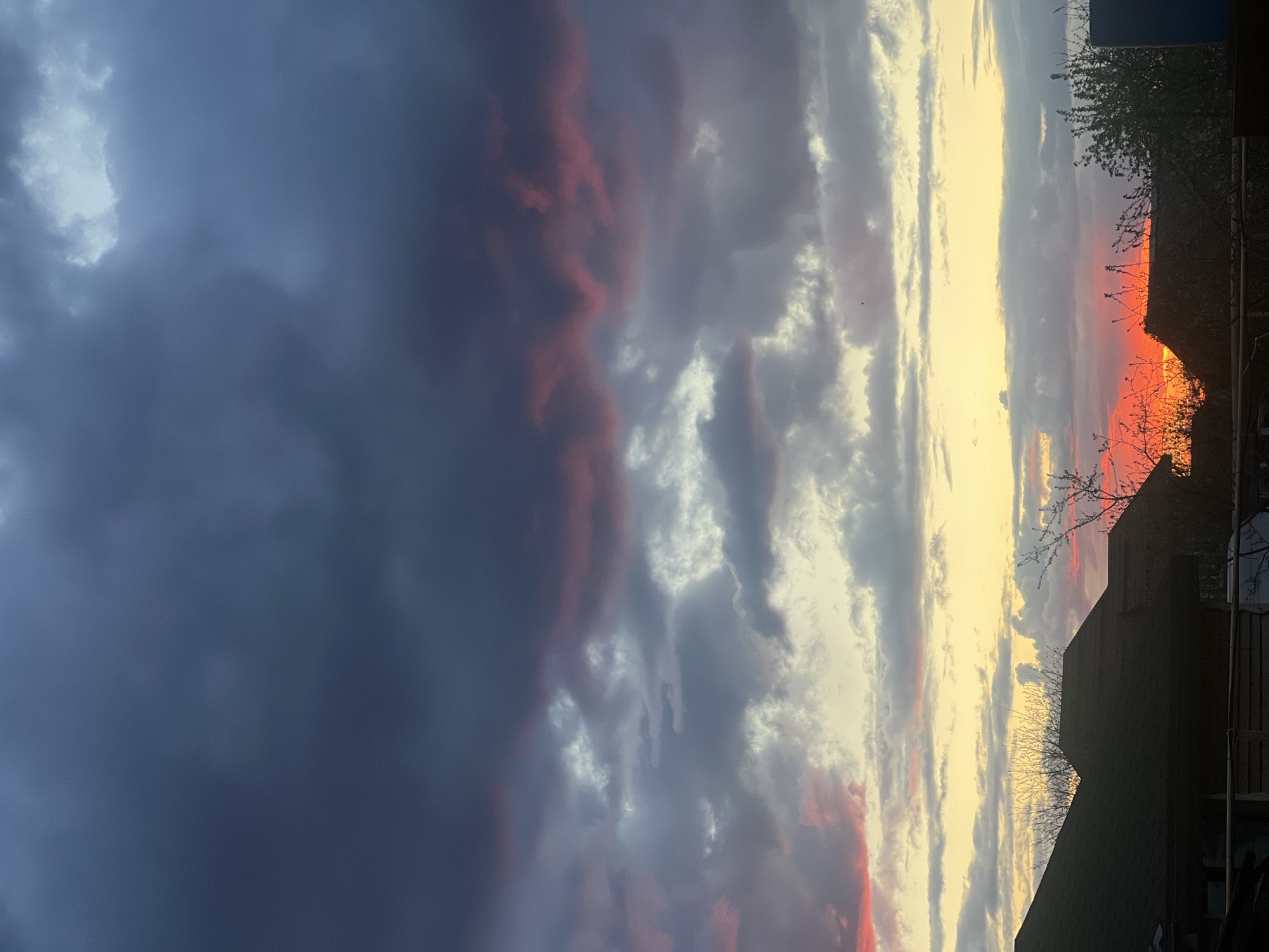 The image depicts a sunset in Votkinsk, Russia. To the left, heavy, dark storm clouds dominate the sky with tinges of red. Moving right, the clouds transition to lighter shades of gray and white, interspersed with patches of red. The right side of the image shows a bright yellow-orange glow, likely from the setting sun. Silhouetted against the sunset are the rooftops of buildings and bare branches of trees.