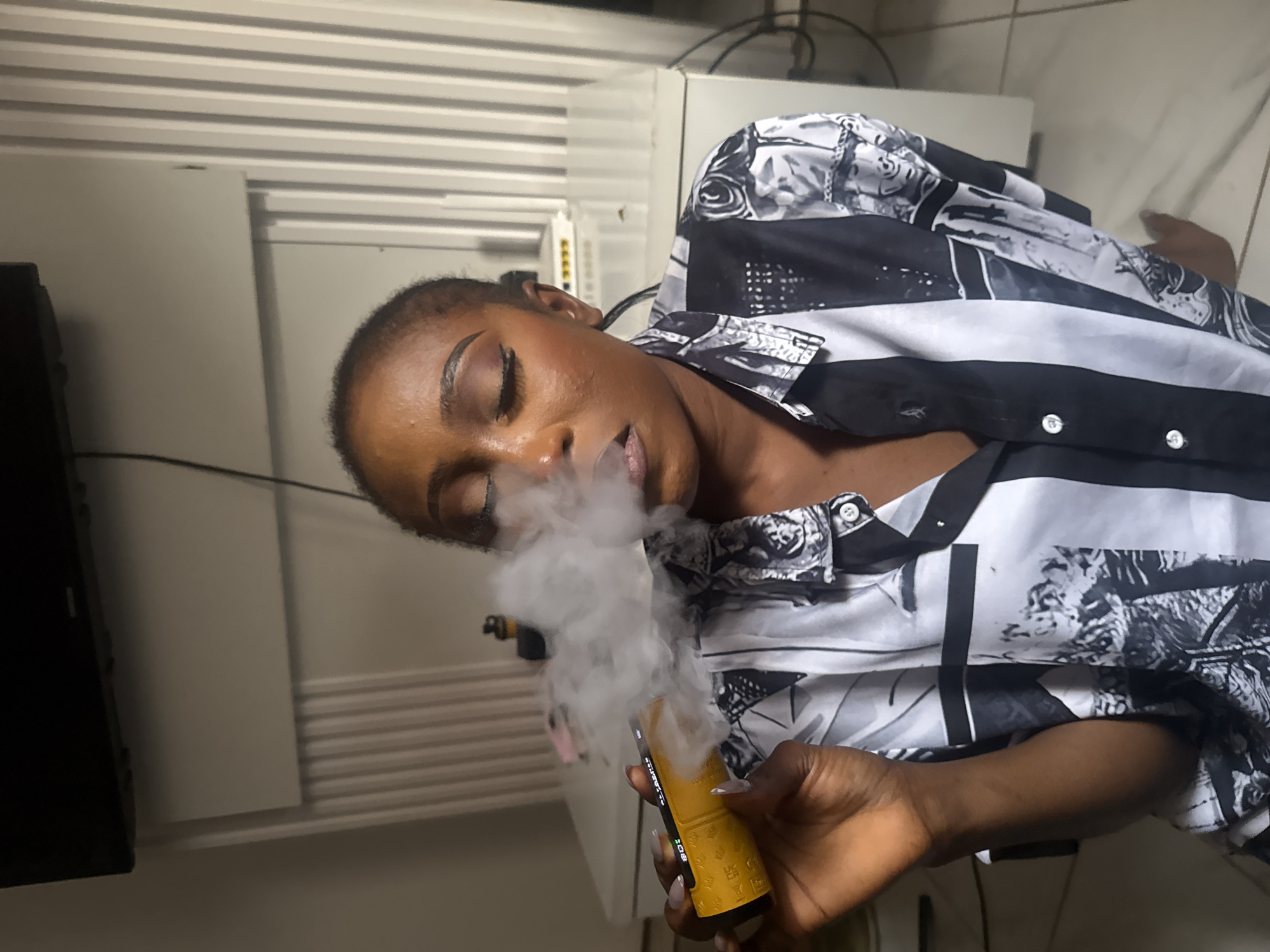 A woman is exhaling a cloud of smoke from a yellow vaping device. She is wearing a black and white patterned button-down shirt and has her eyes closed. The background appears to be an indoor setting with white furniture and a television mounted on the wall. The lighting is soft and diffused, suggesting it may be daytime. The overall mood is relaxed.