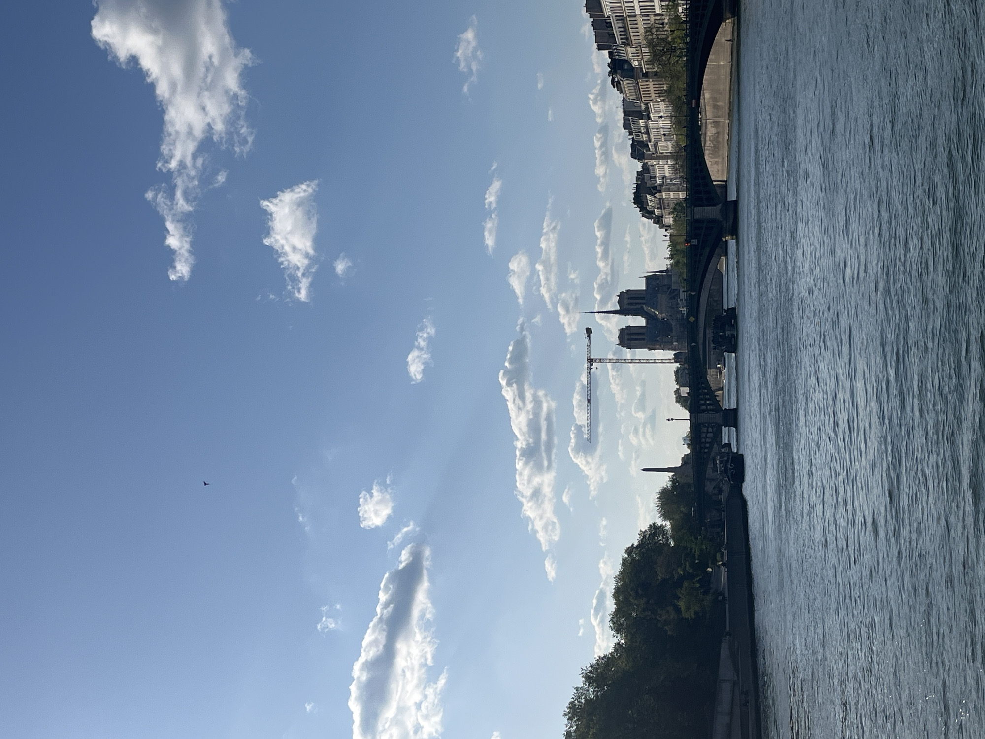 This image captures a wide, sunlit view of the River Seine in Paris on a clear day with scattered white clouds. The sky, a bright blue, fills the upper two-thirds of the frame, adorned with numerous cumulus clouds that reflect the strong sunlight. A small, dark, airplane-like object is visible high in the clear expanse of the sky on the left.
The lower third of the image features the River Seine, its surface showing gentle ripples. The left bank is lined with lush green trees, behind which indistinct urban structures can be seen. The right bank showcases a dense cityscape composed of classic multi-story Parisian buildings.
Prominently featured in the mid-ground, on the right bank, is the iconic silhouette of Notre Dame Cathedral, its distinctive spires and architectural details clearly discernible despite being partially in shadow. Adjacent to Notre Dame, a tall, white construction crane indicates ongoing restoration work. Further left along the right bank, partially obscured by trees, a dark, slender monument resembling an obelisk stands tall. Several bridges span the river, connecting the two banks.
The scene conveys a sense of urban tranquility and historical grandeur under bright, clear weather, likely during the late morning or early afternoon. There is no visible text or identifiable human activity beyond the implicit construction work.