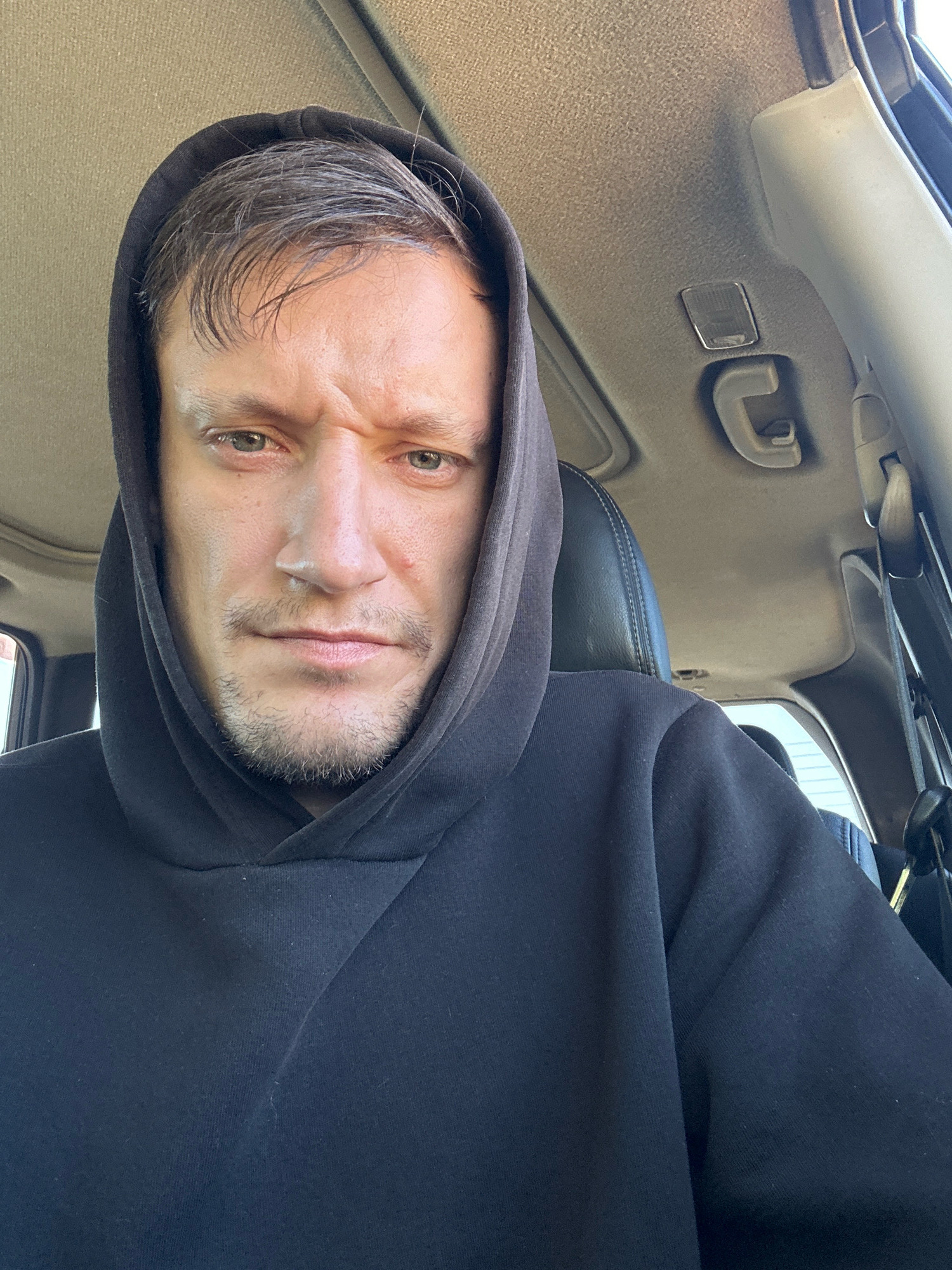 The image is a close-up selfie of a man inside a vehicle, likely a car. The man is wearing a dark hooded sweatshirt, with the hood pulled up around his head. His face is turned towards the camera, with a furrowed brow and a stern expression. The background shows the interior of the vehicle, including the car's ceiling with a sunroof and a grab handle. The lighting suggests it is daytime. The location is not specified in the image itself, and therefore cannot be determined to be the city of Makiv, Ukraine.