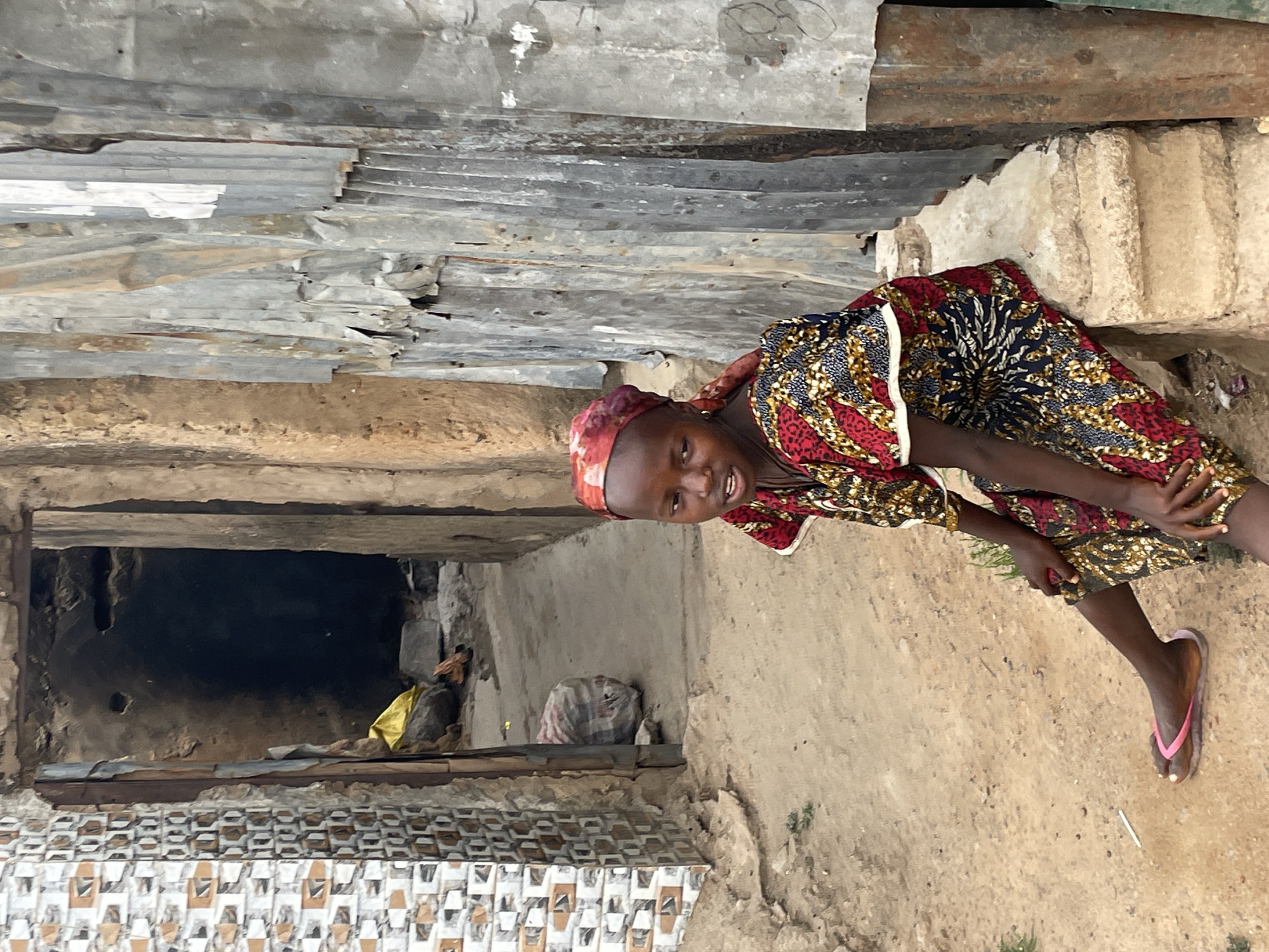 A woman wearing a red and blue patterned dress and a red headscarf is sitting on a concrete step outdoors. She is resting her hand on her leg, wearing flip-flops on her feet, and smiling. Behind her is a corrugated metal overhang and a small, dark opening in a building. The environment appears to be a residential area in Jos, Nigeria. There is a bag and some yellow fabric visible within the opening.