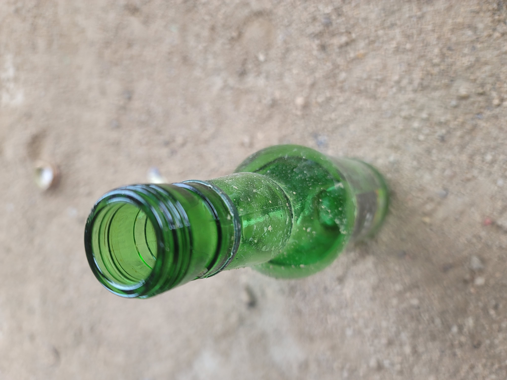 The image shows a close-up, overhead view of a green glass bottle lying on its side on a sandy surface. The bottle is empty and appears to be slightly dusty. The opening of the bottle, which is the most in-focus part of the image, reveals the interior ridges of the neck. The sandy surface is a light brown color with small pebbles and specks of dirt visible. There are no people or any clear indication of a specific activity. The lighting suggests it is daytime. The scene appears to be an outdoor location, possibly a dusty pathway or ground area, given the sandy texture and scattered debris. The image is a still photograph with no discernible text.