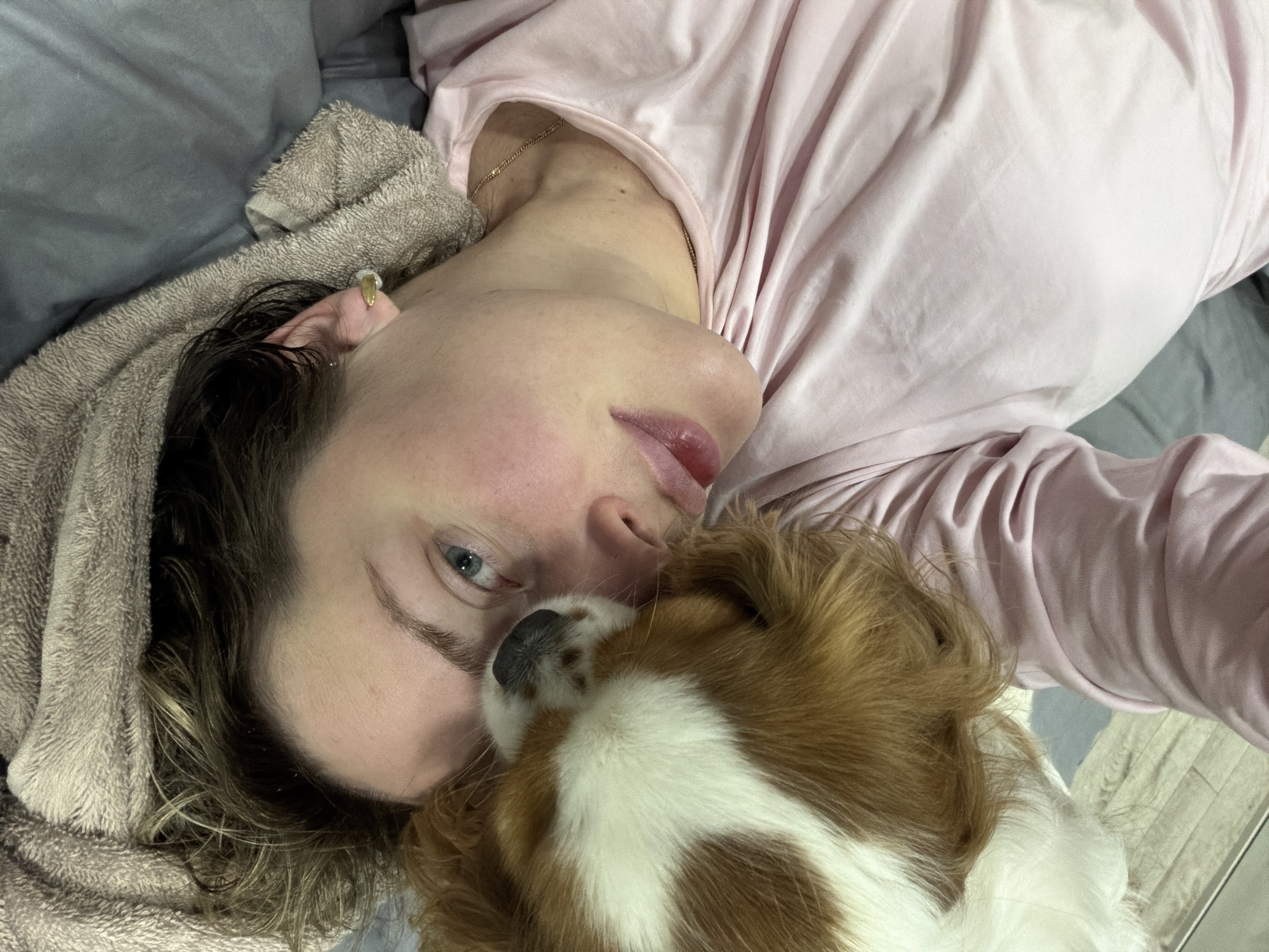 The image captures a close-up, top-down perspective of a person and a small dog in an intimate indoor setting. A person, appearing to be a woman, lies on her back, her head partially resting on a beige, textured towel or blanket. She is wearing a light pink top, and a thin gold chain necklace is visible around her neck. A gold earring adorns her ear. Her eyes are open, looking towards the viewer with a calm, neutral expression. Her dark, wavy hair is visible around her head.
A small dog, with distinct patches of brown and white fur, is snuggled closely beside the person's face, its dark nose touching her cheek. This suggests a moment of affection or shared rest between the two. The background features gray bedding or sheets, with a small section of light-colored flooring visible in the bottom right. The scene conveys a quiet, relaxed atmosphere of companionship. No specific time of day or external weather conditions are indicated, and no text is visible in the image.