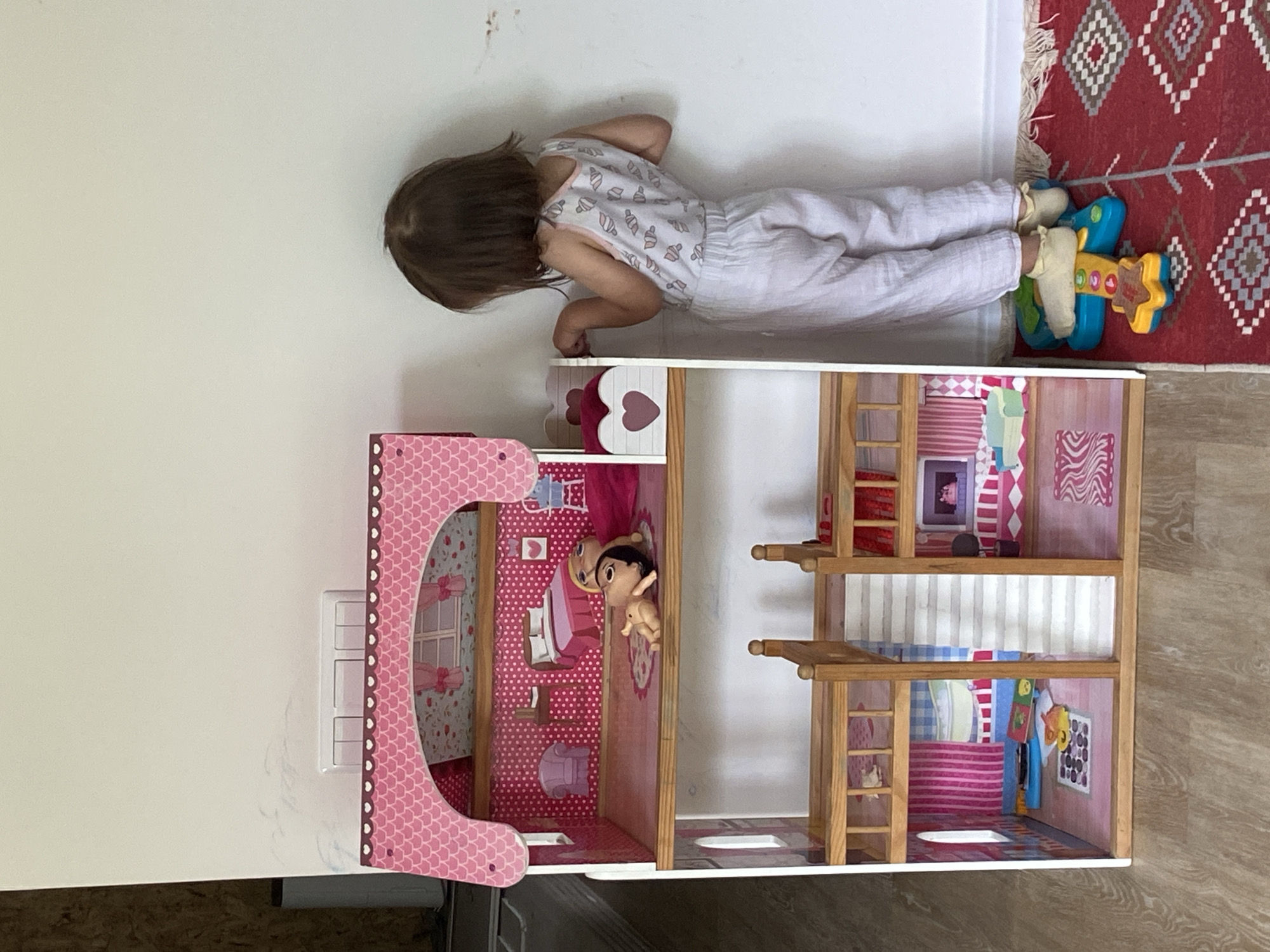 A young child, lying prone on a cream-colored surface, is playing with a dollhouse. The child, wearing a patterned white onesie, is positioned partially over the dollhouse, which is multi-story and features detailed interior decor in shades of pink, white, and blue. The child's head is closest to the top edge of the image, and their feet, which are near a red patterned rug, are closest to the right edge. The dollhouse is adjacent to a white wall. The location is Tyumen, Russia.