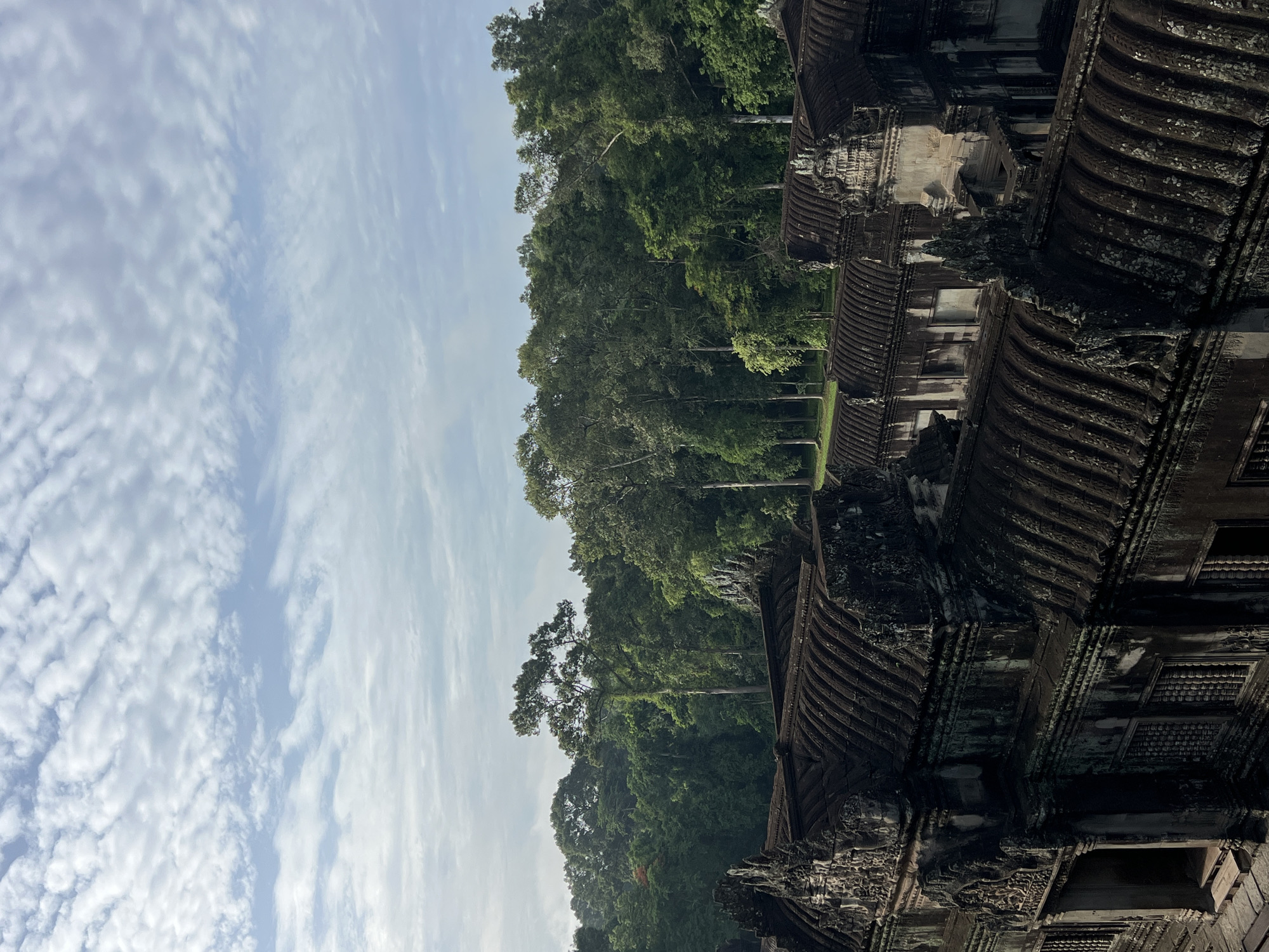 The image shows an aerial view of a large, ancient stone temple complex in a jungle environment. The foreground is dominated by the weathered, dark stone structures of the temple, featuring intricate carvings and tiered roofs. The architecture suggests a historical significance, possibly religious in nature. Beyond the temple, a dense, lush green forest stretches to the horizon, with tall trees and a thick canopy. The sky above is partly cloudy, with wispy white clouds scattered across a pale blue expanse. The overall impression is one of age, grandeur, and a harmonious integration with the natural landscape. The location is identified as Angkor Wat, Siem Reap, Cambodia.