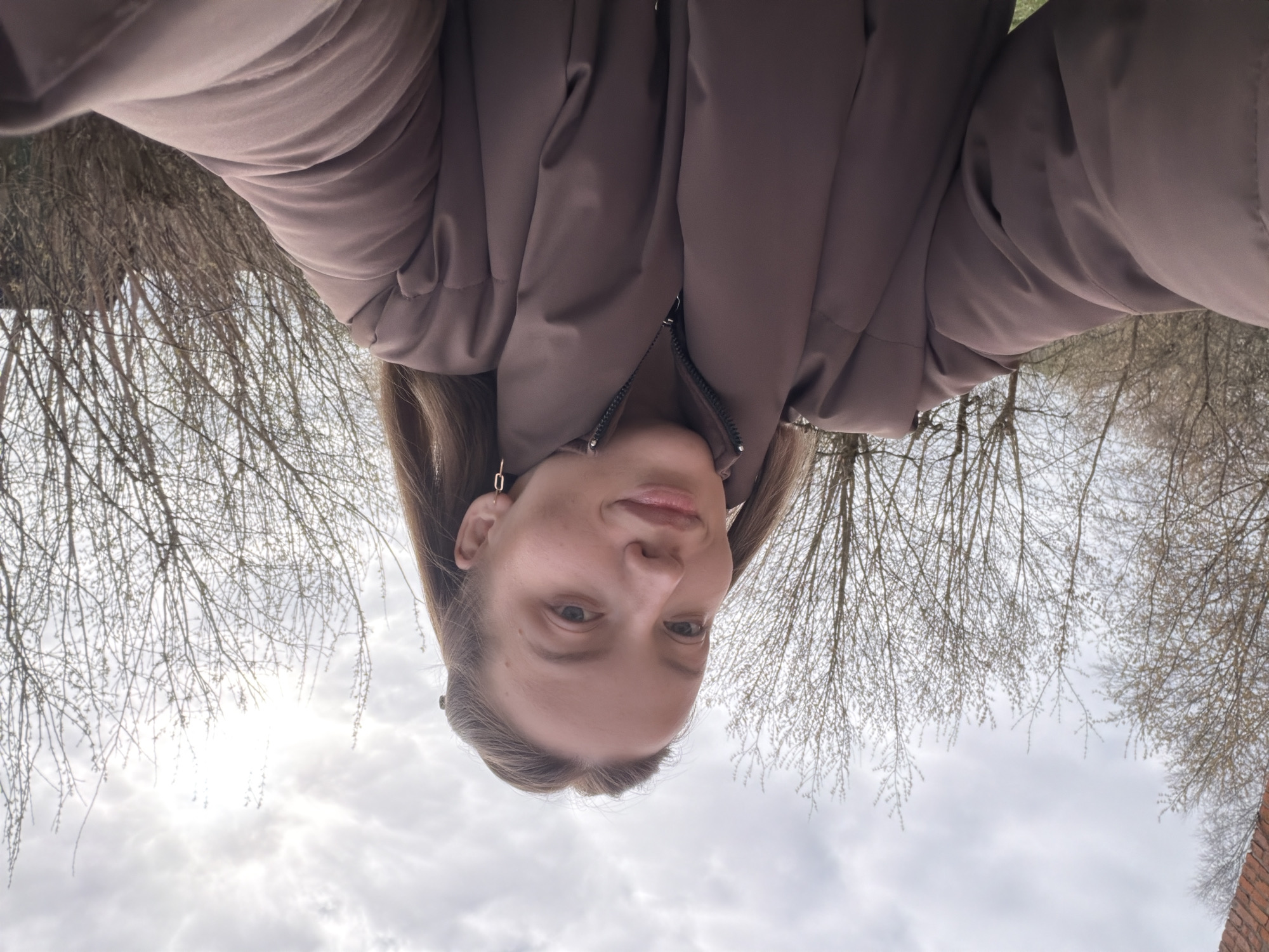 A woman wearing a brown puffer jacket is depicted in a selfie taken outdoors. Bare trees are visible in the background. The sky is overcast. The location is likely Reshetnikovo, Russia.