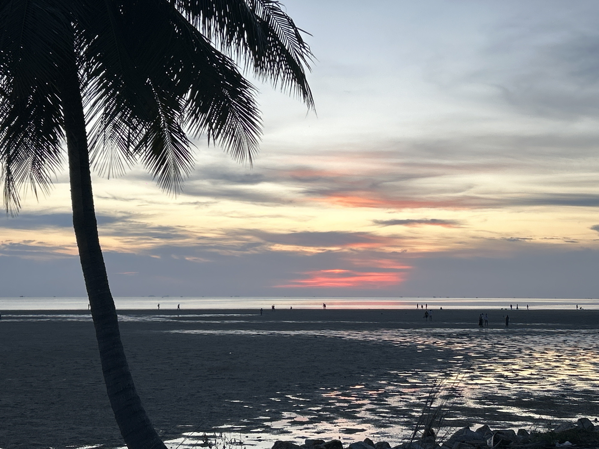 The image captures a twilight scene at a beach in Baan Sri Thanu, Thailand. A silhouette of a palm tree dominates the left foreground. In the background, the sky is a blend of muted oranges, pinks, and grays, suggesting either sunrise or sunset. Below the horizon line, a shallow, tidal flat extends toward the sea. Several individuals are sparsely distributed across the flat, appearing as silhouettes against the water. The foreground shows a slightly wet beach with rocks and some discarded debris.