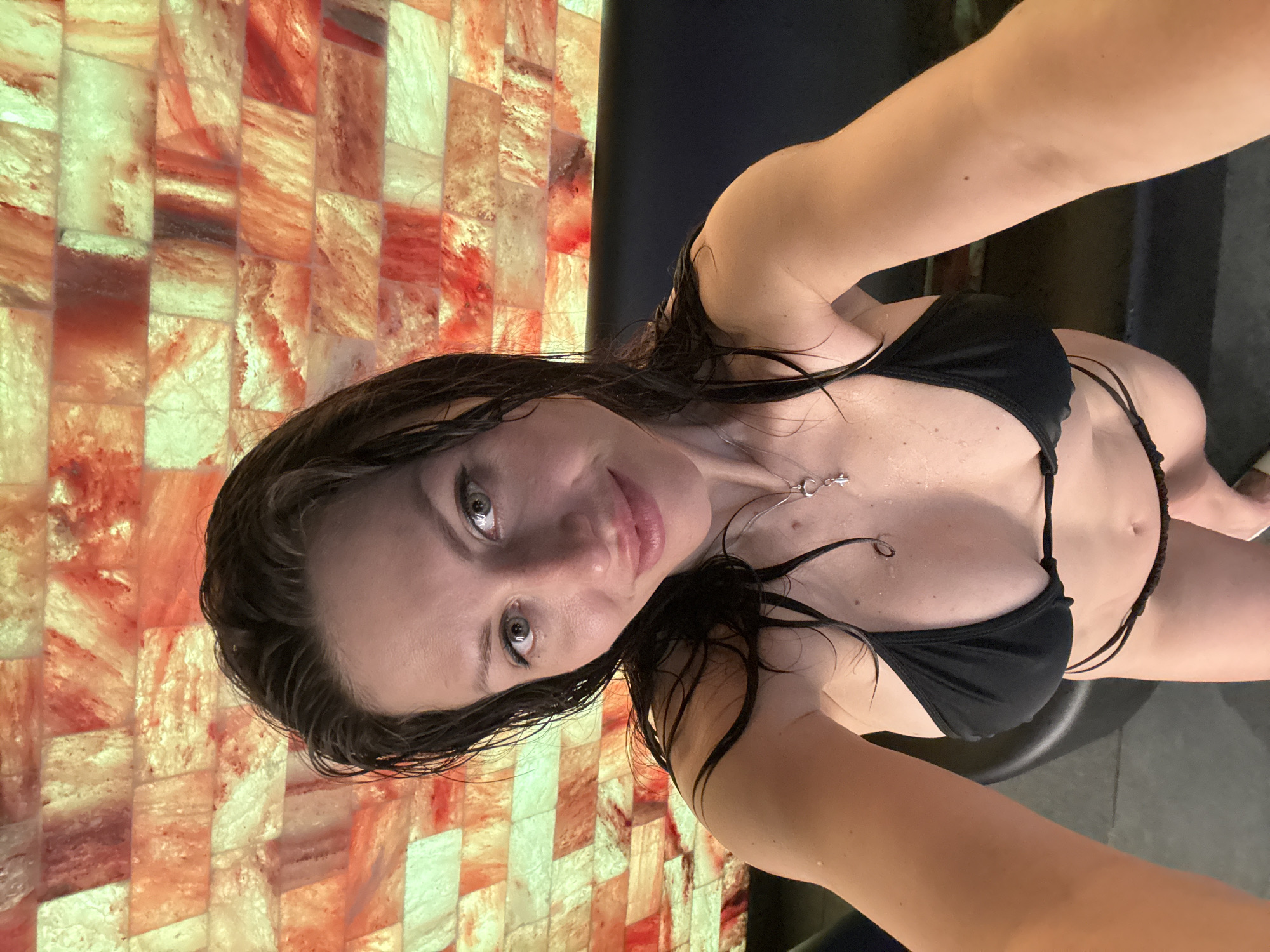 A woman with wet brown hair is taking a selfie while wearing a black bikini. She has light eyes and fair skin. A silver necklace is visible. She is standing near a wall constructed of illuminated salt blocks. The setting appears to be indoors, possibly a spa or sauna. There is no discernible information to indicate that the picture was taken in Saint Petersburg, Russia.