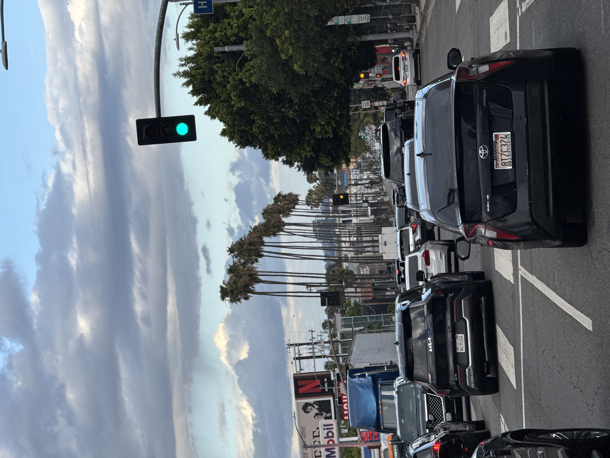 The image shows a street scene in Los Angeles, United States of America, characterized by heavy traffic congestion. The view is oriented along a street lined with palm trees. Several vehicles are visible, including a black Toyota Prius with California license plate "8TZC324" and a black Kia SUV with license plate "8635164". A traffic light is illuminated with a green signal. A Mobil gas station sign is visible in the lower left, with fuel prices displayed. Overcast sky and clouds are above, with hints of blue sky.