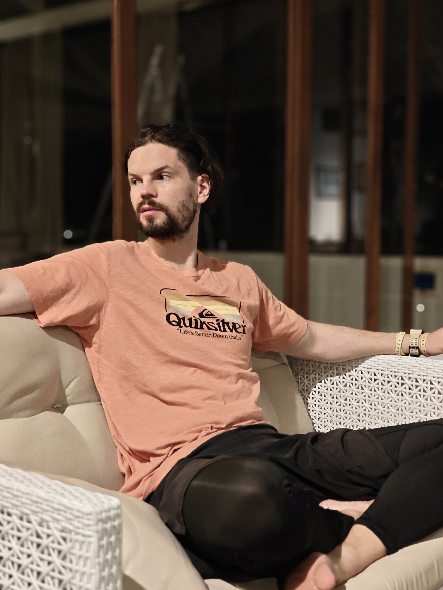 A man is seated on a light-colored, woven armchair in an indoor setting. He is wearing a peach-colored t-shirt with a "Quiksilver" logo and black pants. He has dark hair and a beard and is looking off to the left of the frame. He is wearing several wristbands or bracelets on his right wrist. The background shows an interior space with a window or glass panel featuring dark wood framing. The exact location is Kalote Mokashi, India.