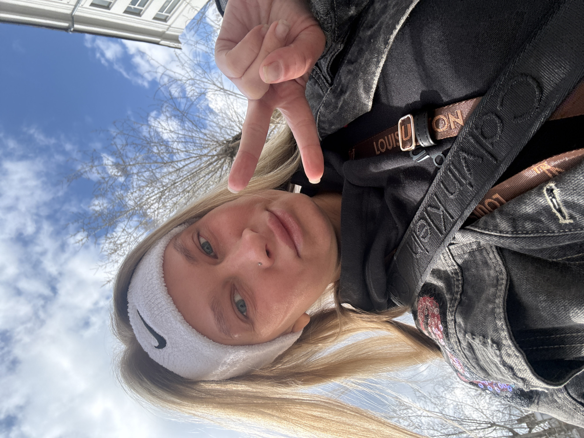 A young, fair-skinned woman with blond hair and blue eyes is shown in a selfie taken outdoors. She is wearing a white Nike headband and a black hooded jacket with a Louis Vuitton strap across her chest. She is making a peace sign with her hand. The background includes a building and a blue sky with clouds. Based on visual cues, it is impossible to discern the city of Perm, Russia, or other specific location details.