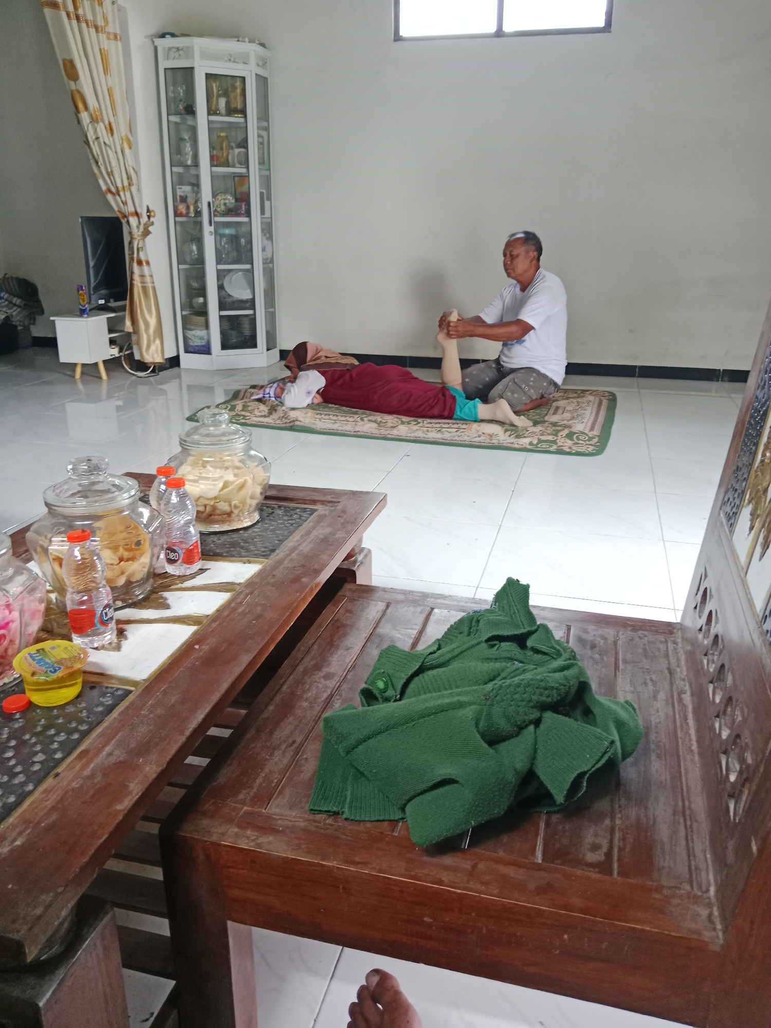 In a room in Mangunan, Indonesia, a man kneels on a mat, performing a foot massage on a woman who is lying on her back on the same mat. The woman is wearing a maroon headscarf and a blue garment. The man is wearing a white t-shirt and patterned shorts. To the left of the massage scene, a wooden coffee table is laden with glass jars filled with snacks, plastic bottles of water, and a yellow tub. In the background, a tall, white glass-fronted cabinet displays various items, and a television is visible on a small white stand next to a cream-colored curtain. A dark green knitted garment is draped over a wooden chair in the foreground, and a portion of a bare foot is visible at the bottom of the frame.