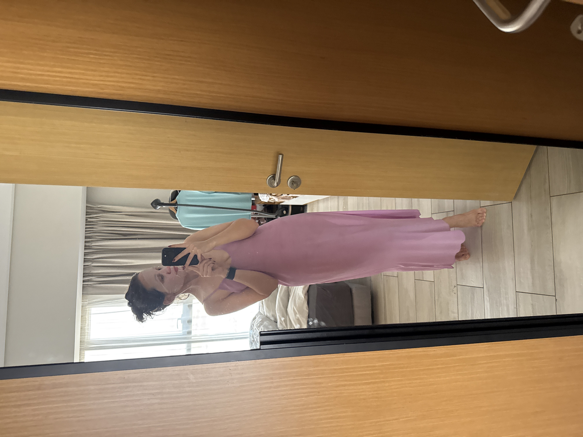 A woman is reflected in a full-length mirror, possibly in a hotel room in Dubai, United Arab Emirates. She is holding a smartphone up to take a picture of herself. The woman has dark hair and is wearing a long, light pink dress. She is standing on a tile floor. Behind her, in the reflection, are a bed and window with light coming in. A turquoise garment is hanging in the background as well. The mirror is set into a wooden frame. A door handle is visible at the top of the image.